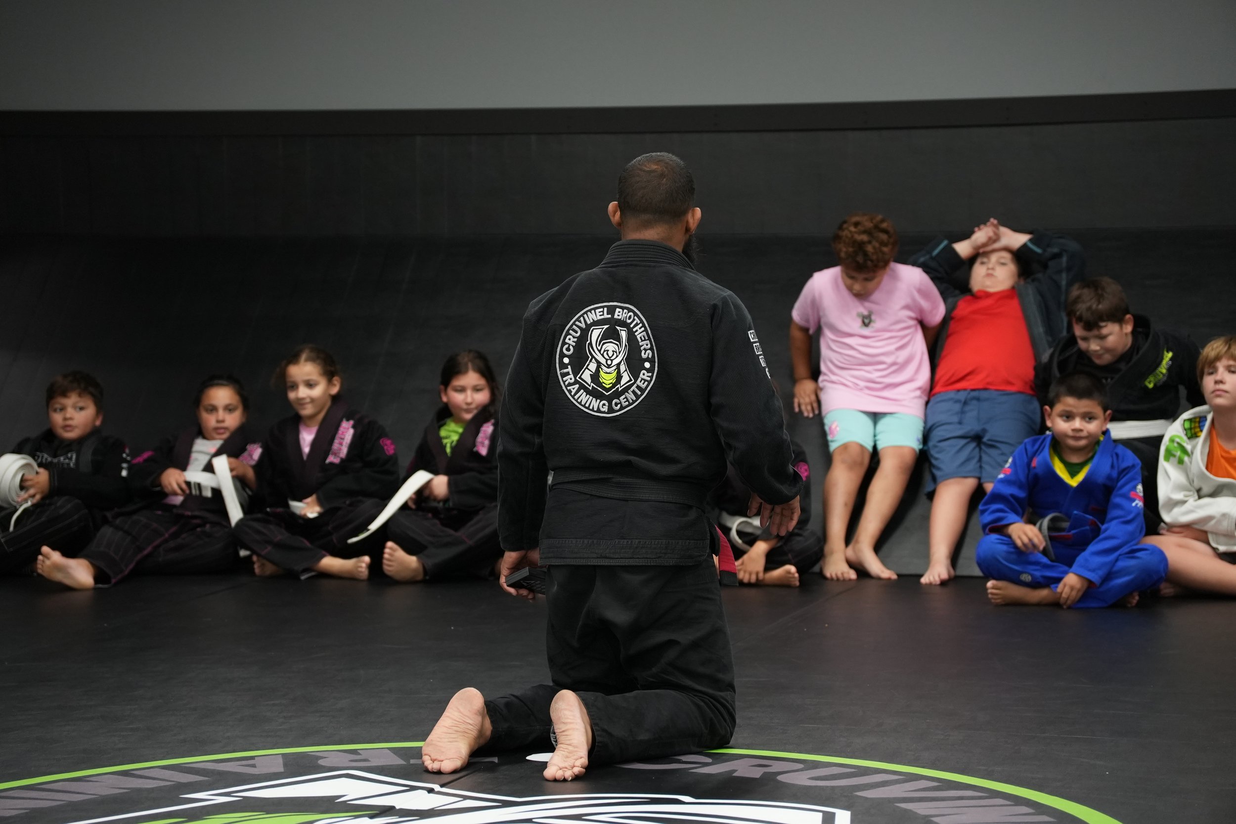 Martial arts instructor in a black gi with patches on the back, teaching a class of children sitting on the mat, some in gi uniforms and others in casual clothing, in a training center.