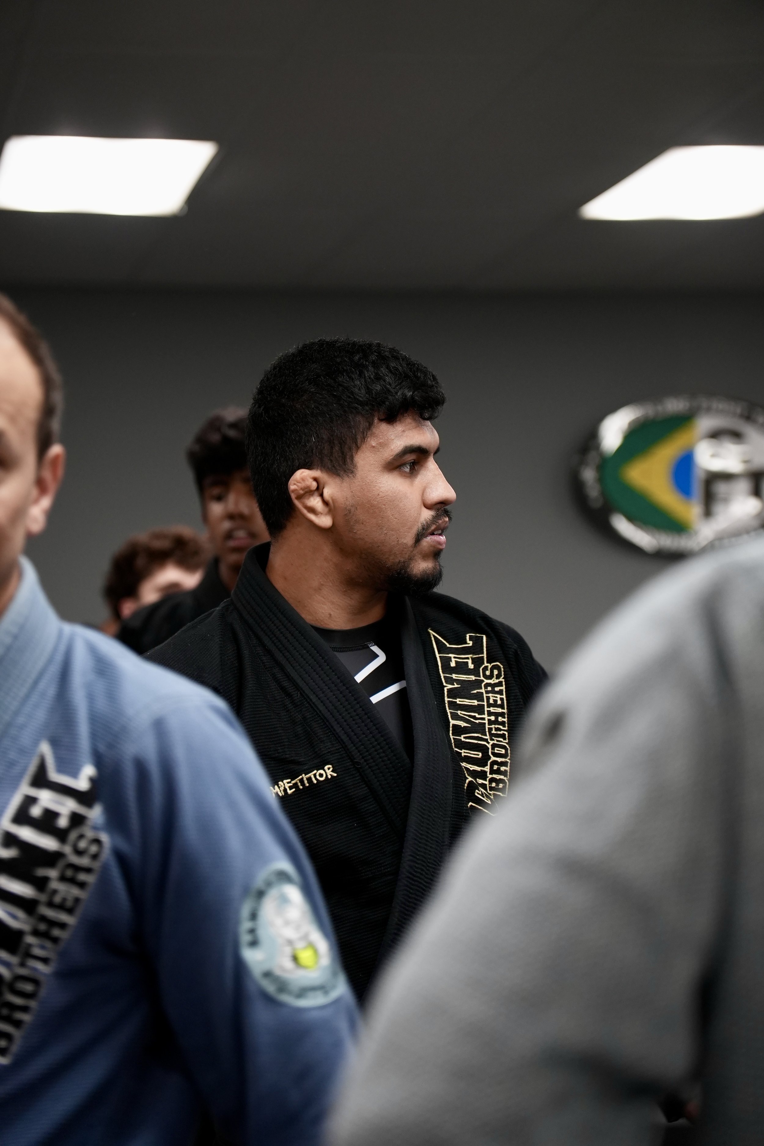 A man with dark hair and a beard wearing a black Brazilian Jiu-Jitsu gi with a gold sponsor patch standing in a martial arts training facility, surrounded by other practitioners.