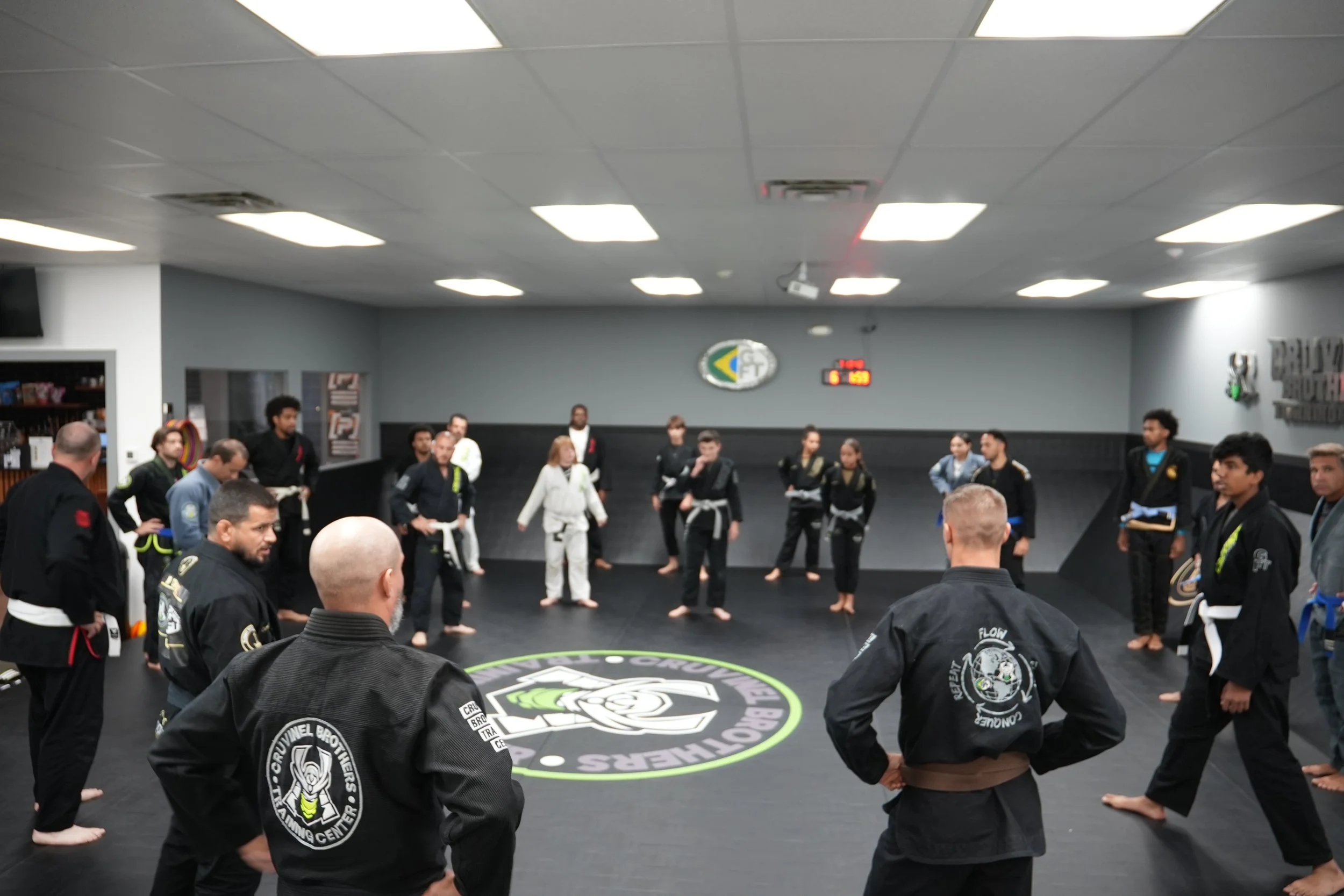 Group of martial arts students and instructors standing in a circle in a gym, wearing traditional uniforms with patches, inside a martial arts school.