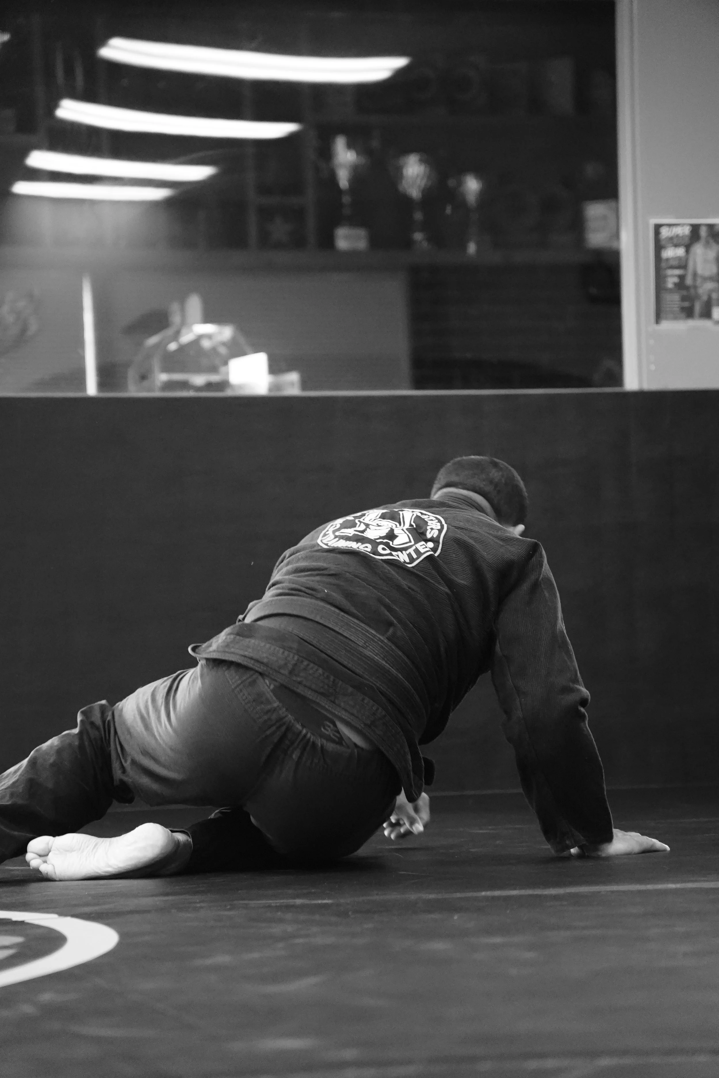 A person practicing Brazilian Jiu-Jitsu on a mat, wearing a gi, with a focus on the seated position and facing away from the camera.
