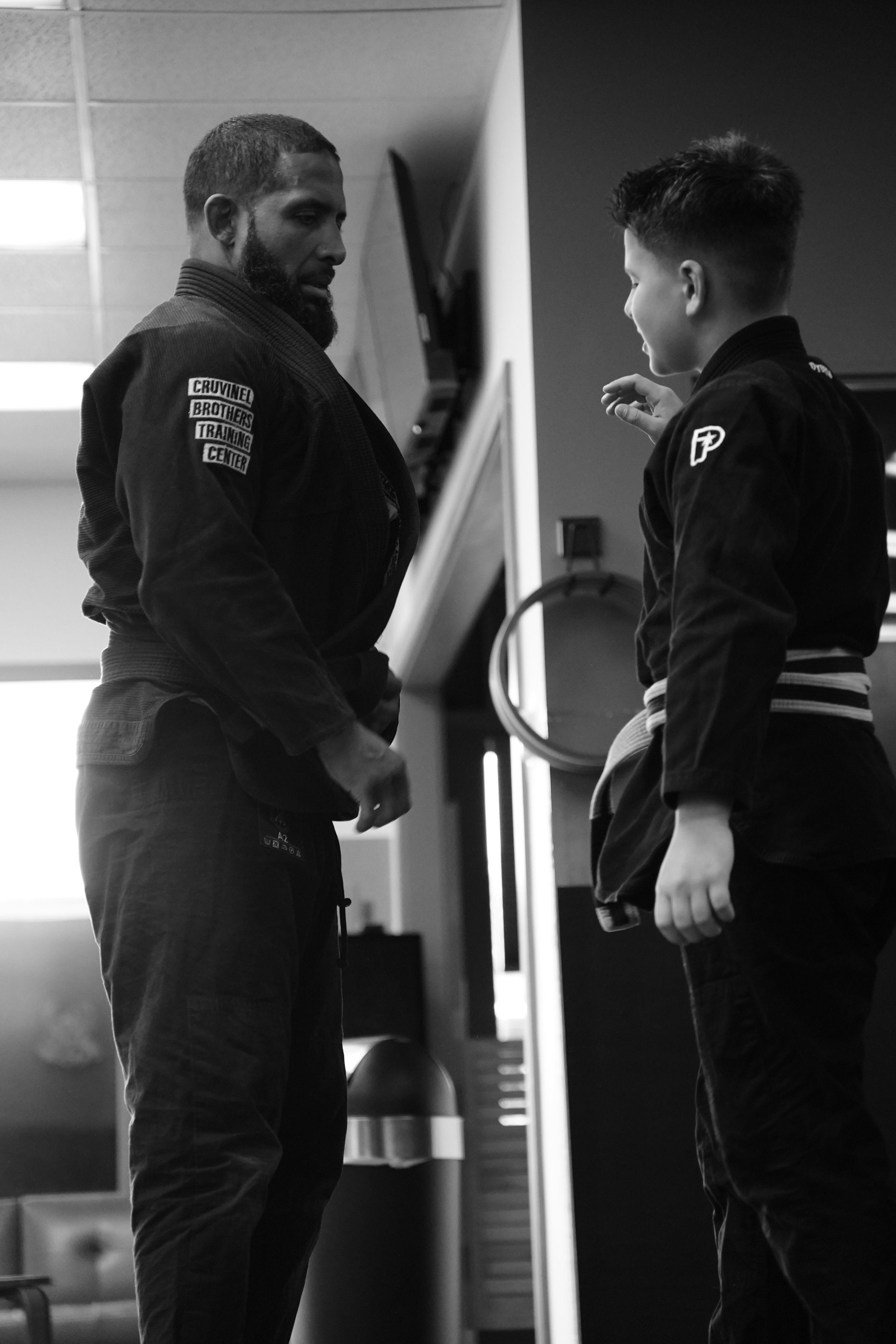 A black and white photo of a martial arts instructor and a young student in a gym, wearing traditional gi uniforms with patches, standing and talking.