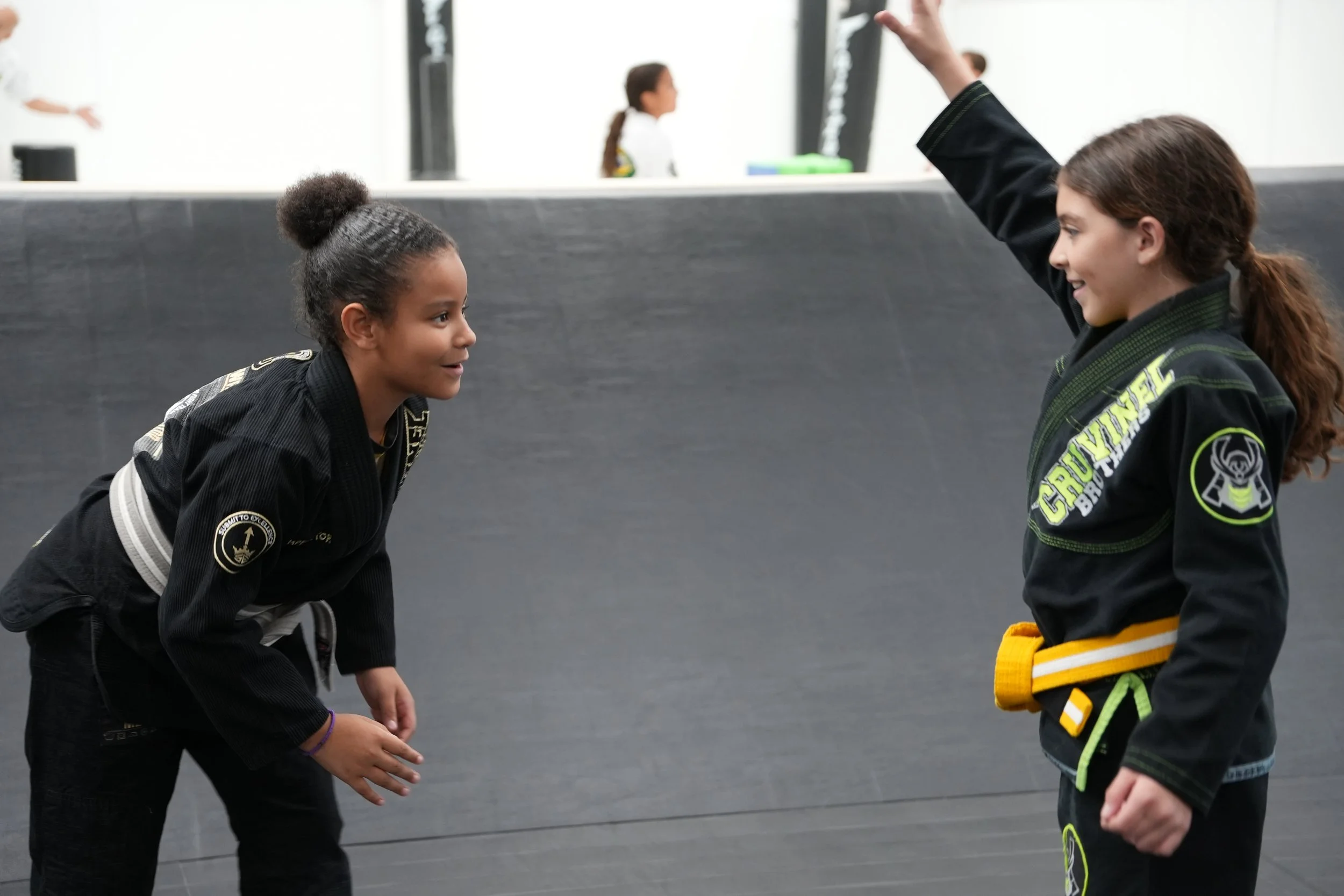 Two young girls in Brazilian Jiu-Jitsu uniforms engaging in a training session, with one girl appearing to instruct the other, on a black mat in a gym.