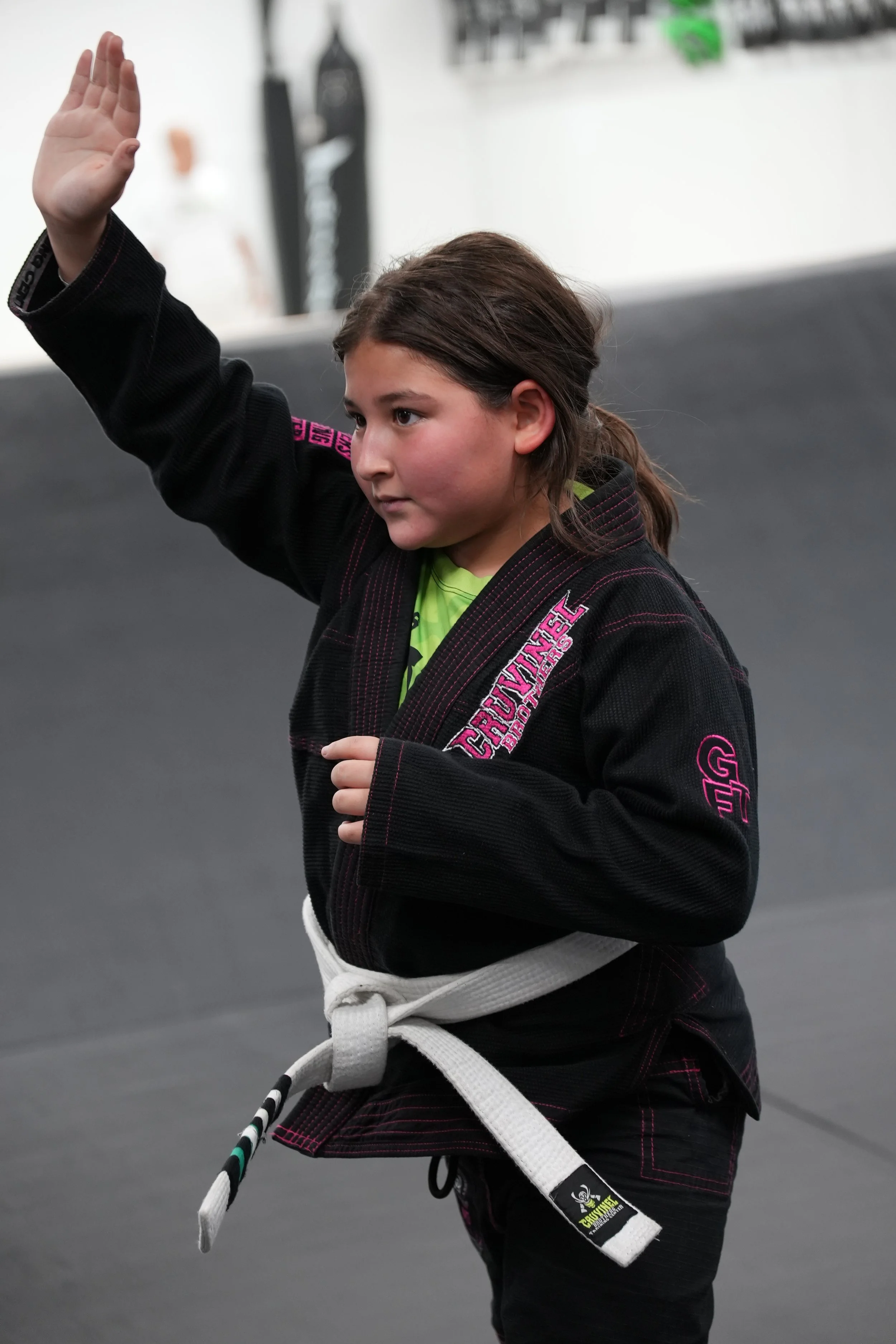 Young girl in a black martial arts gi with pink and green accents practicing a martial arts stance in a dojo with black mats, holding a black and white martial arts weapon.