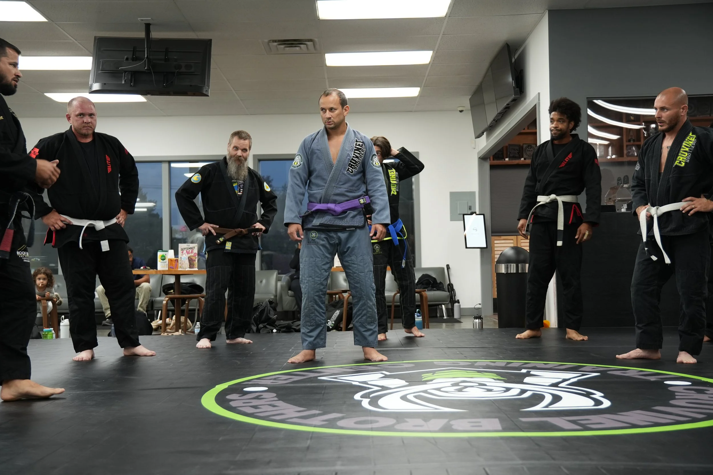Martial arts students and instructor in a dojo, wearing gis and belts, gathered on a black mat with a circular logo, during a training session or class.