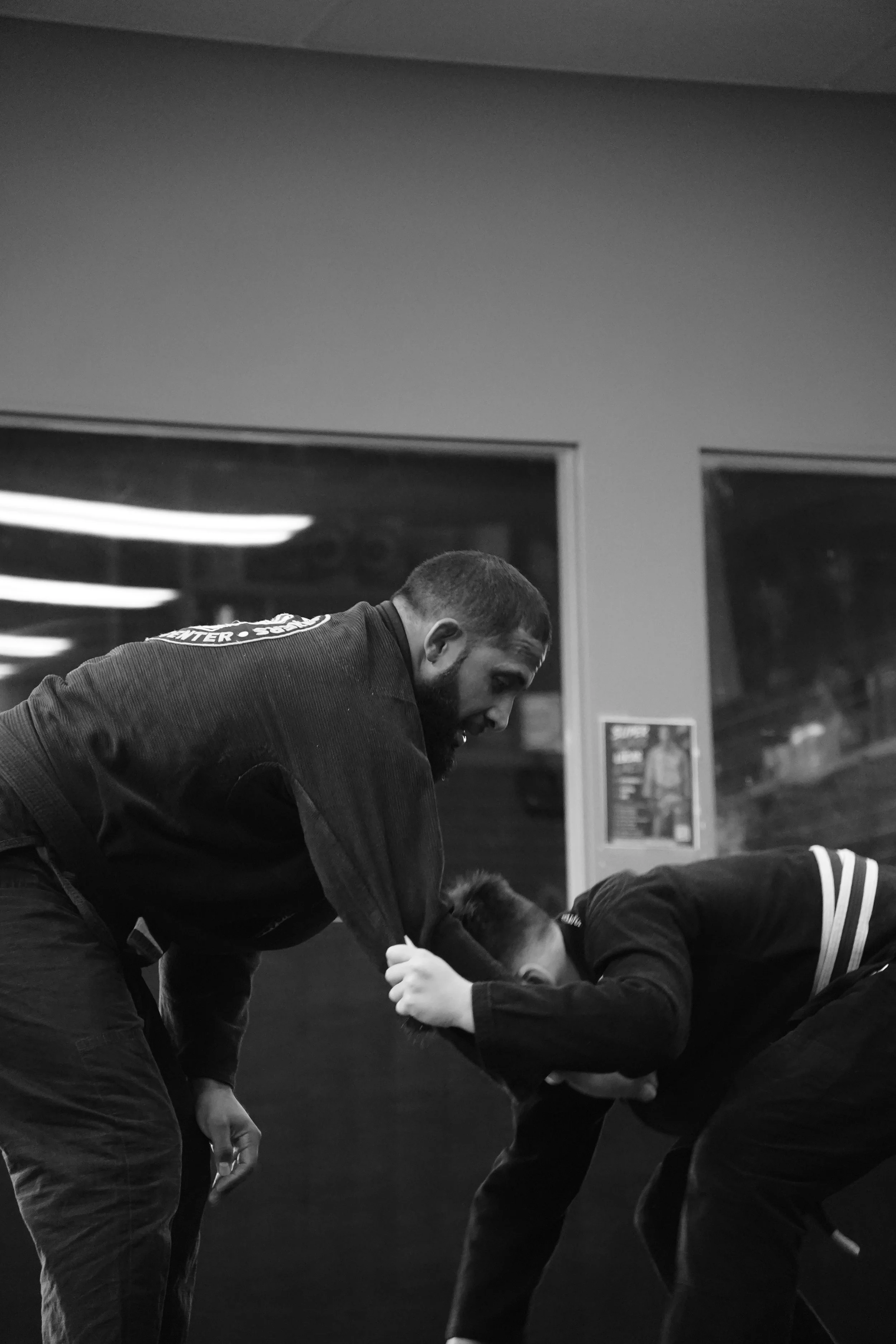 A man in martial arts training gear bending over to assist a young martial artist during practice or competition in a gym or dojo, with posters and windows in the background.