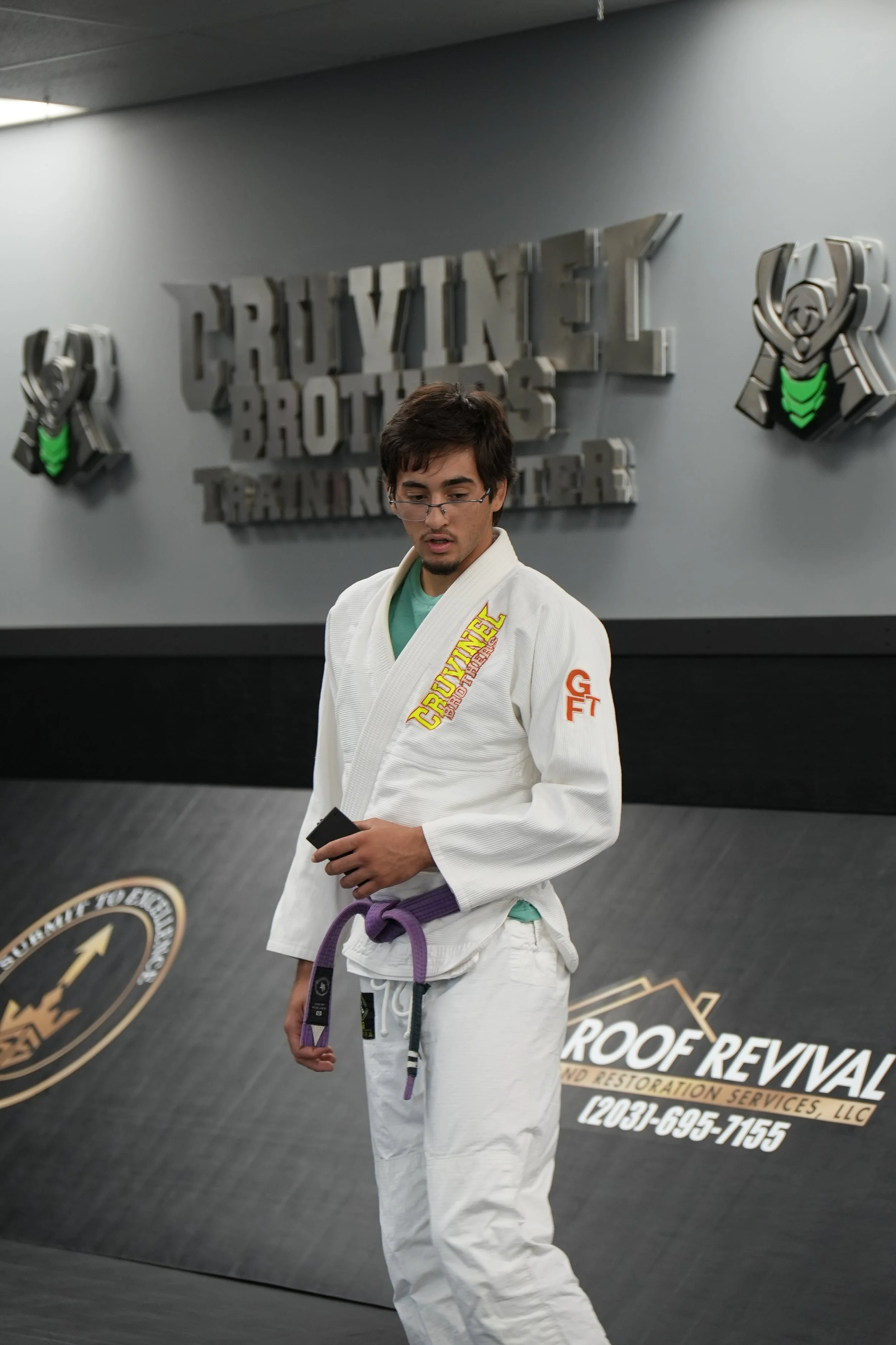 A man wearing a white Brazilian jiu-jitsu gi with purple belt standing in a gym with gray walls, and a sign that reads 'Roof Rehab' and other business details.