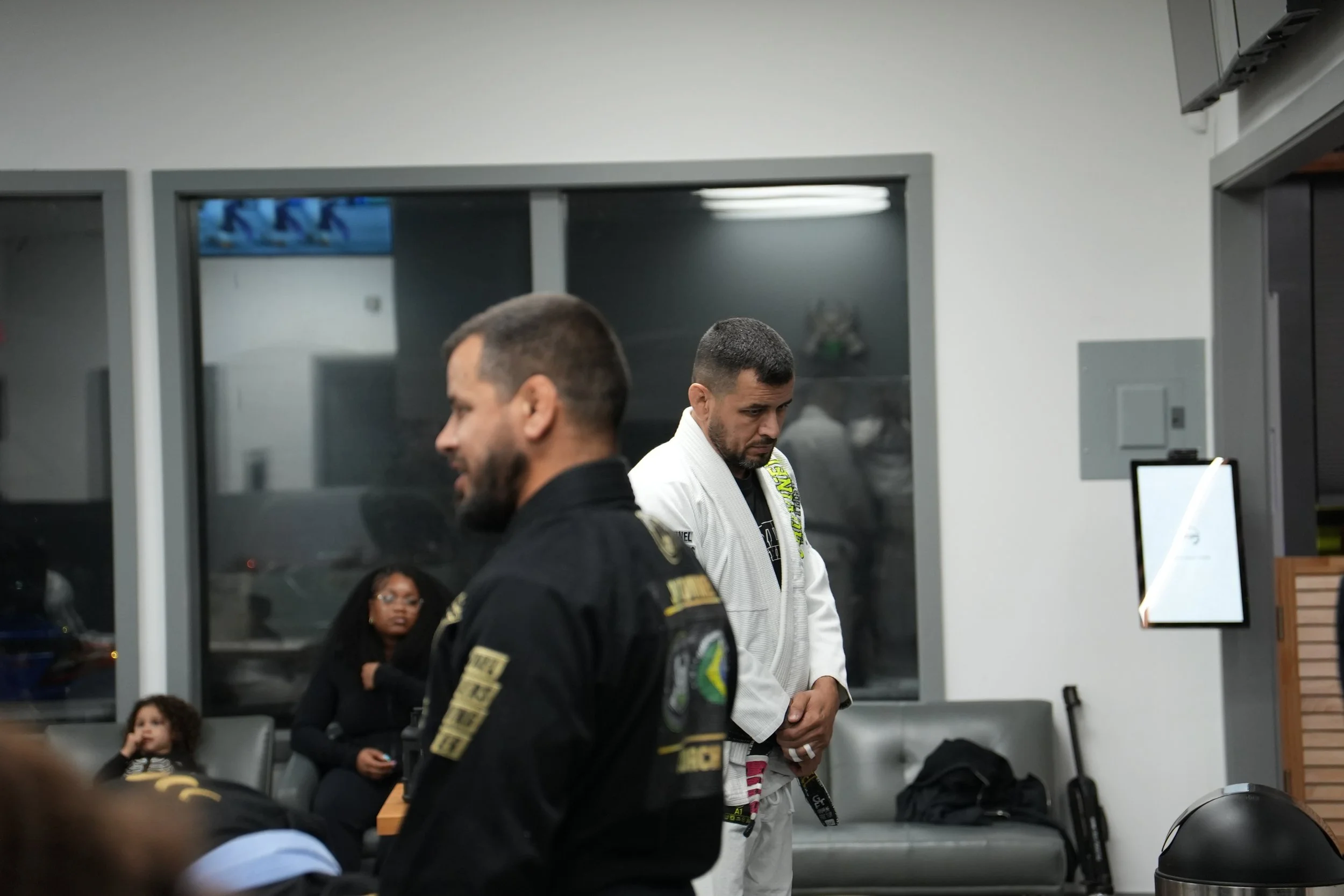 Inside an airport or transportation hub, there are several individuals including two men in focus wearing Brazilian Jiu-Jitsu uniforms and patches. A woman and a child are seated behind them, with the woman looking at something, and the child observi