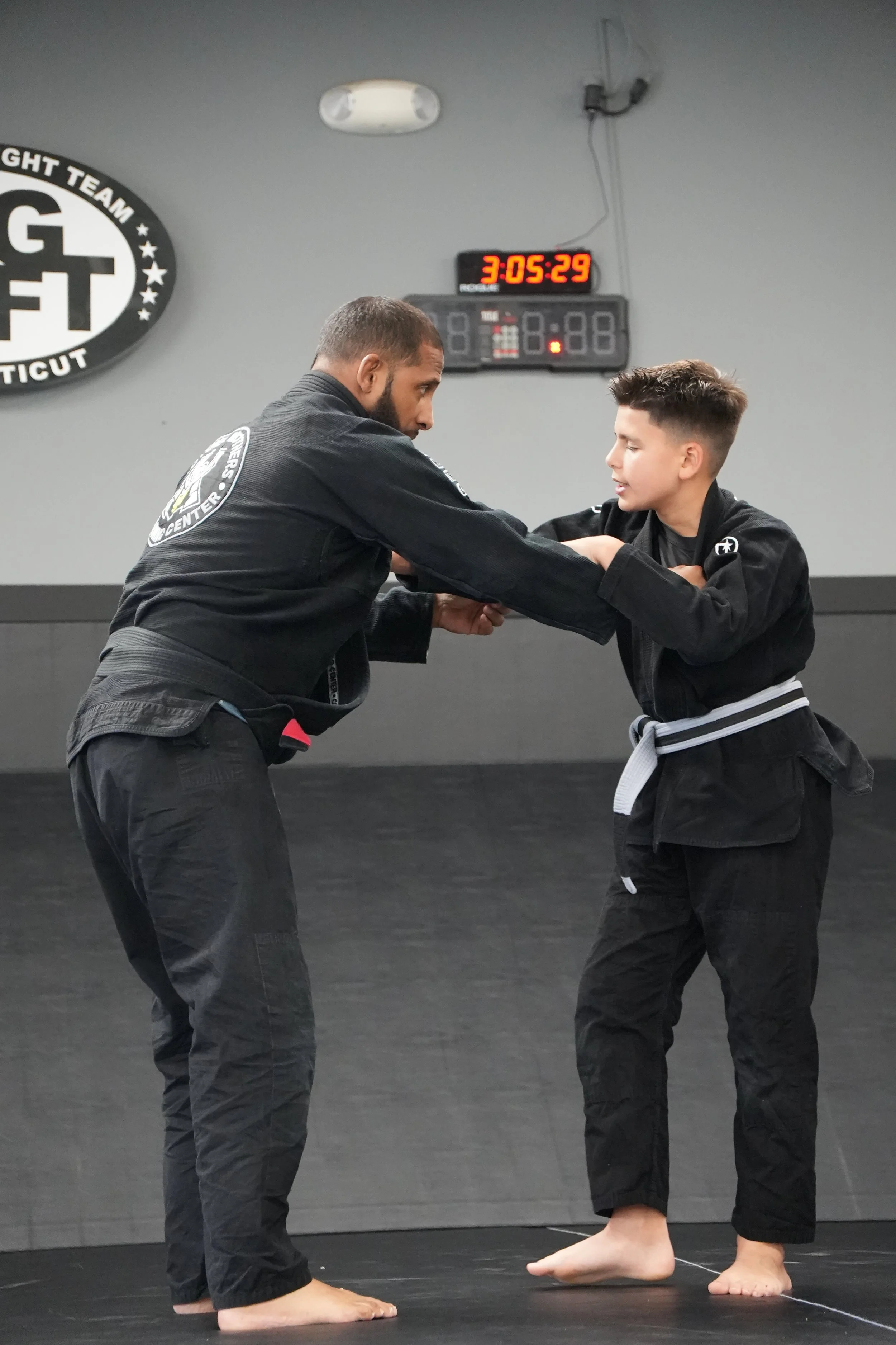 Two martial artists practicing Brazilian Jiu-Jitsu in a training gym. An adult man and a teenager are wearing black gi uniforms, standing on a black mat, engaging in a grappling position with their hands clasped. The man is taller, with a short beard