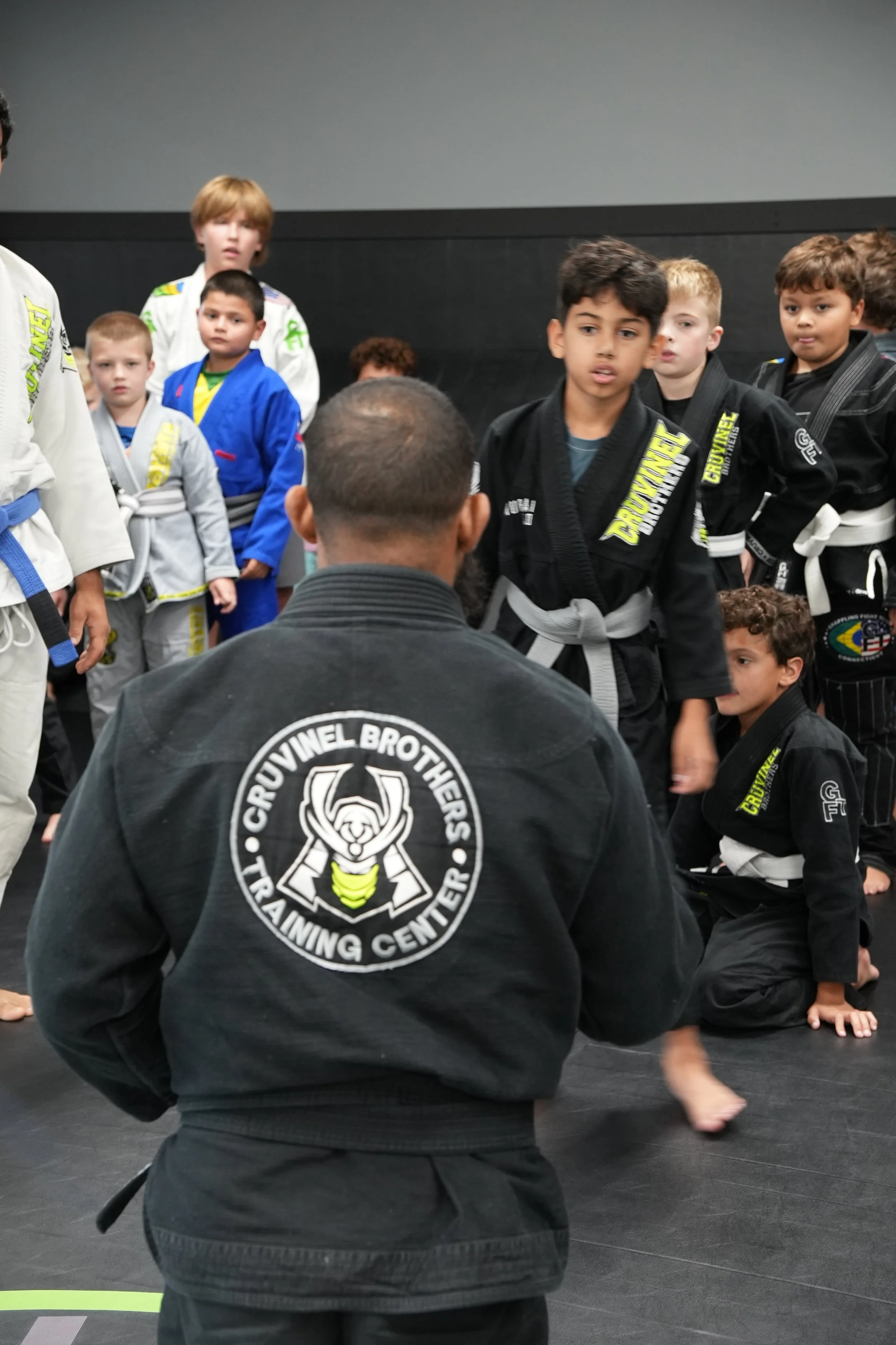 Brazilian Jiu-Jitsu students in uniform indoors, listening to instructor, with various colored belts, some standing and some kneeling on the mat.
