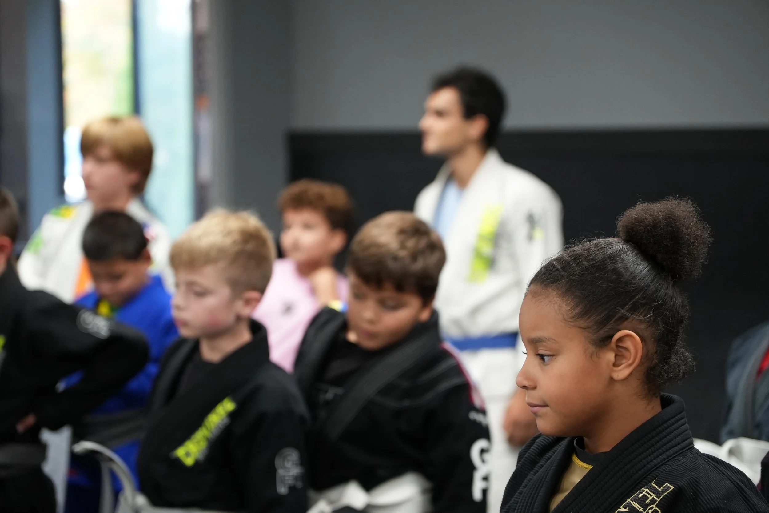 Group of children and young adults in a martial arts class, with some wearing black uniforms, in a gym or classroom setting.