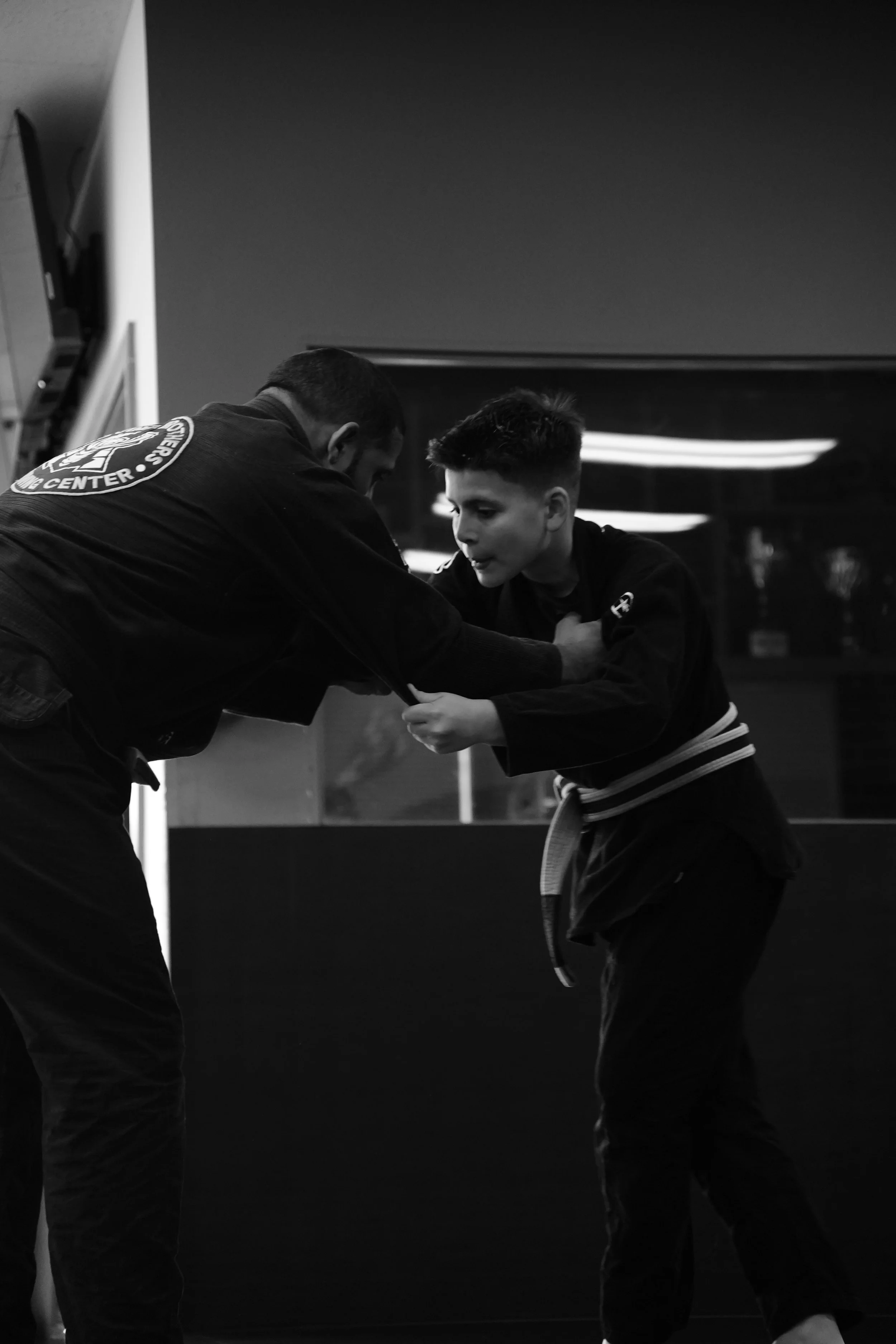 Two males practicing Brazilian Jiu-Jitsu, wearing gi uniforms, engaged in a grappling move in a dojo. One appears to be instructing or assisting the other.