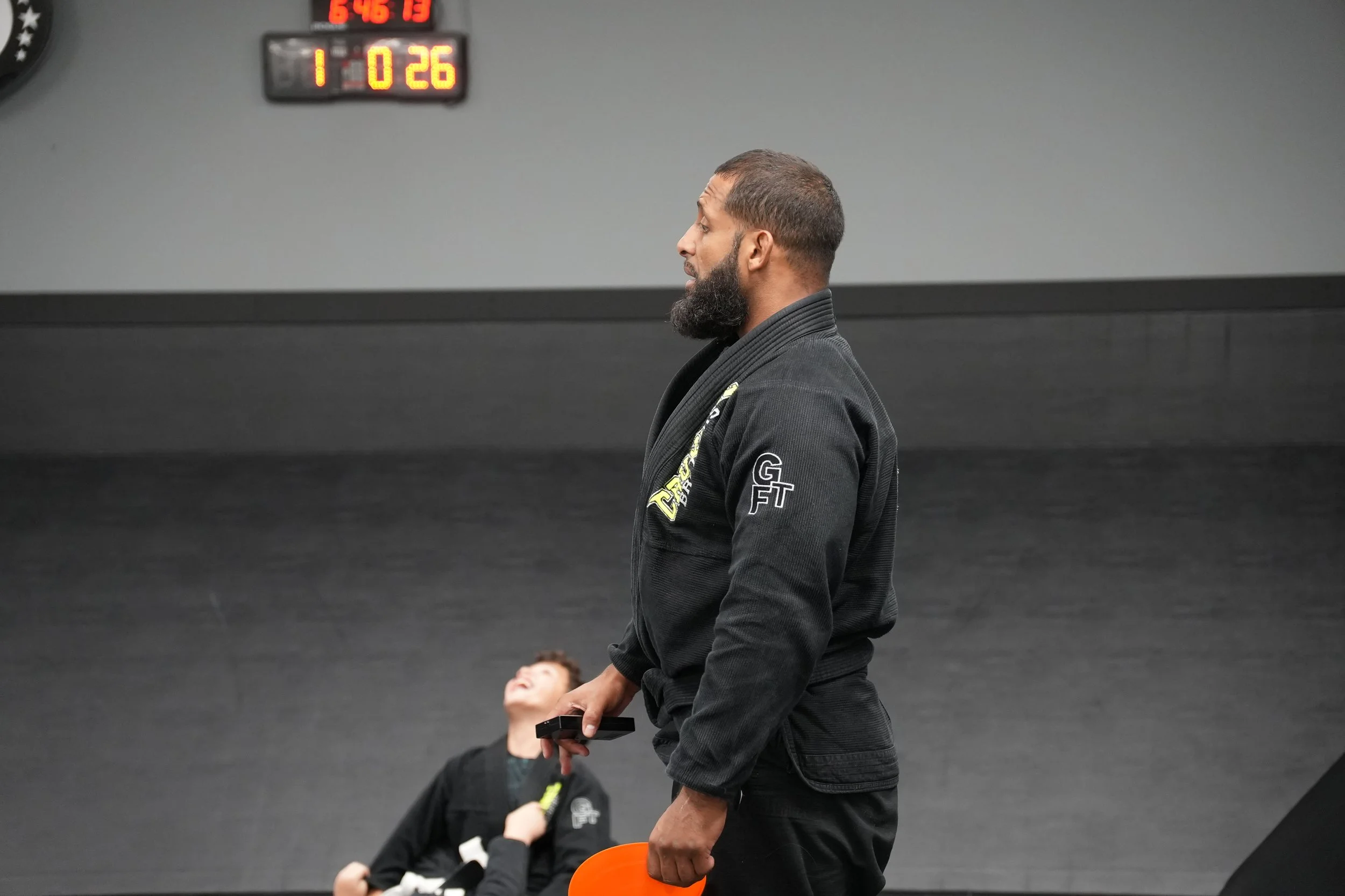 A man with a beard and short hair wearing a black martial arts gi shirt standing in a black martial arts gym, holding a small black rod and an orange object, with children in the background, one of whom is looking up at him. There is a digital scoreb