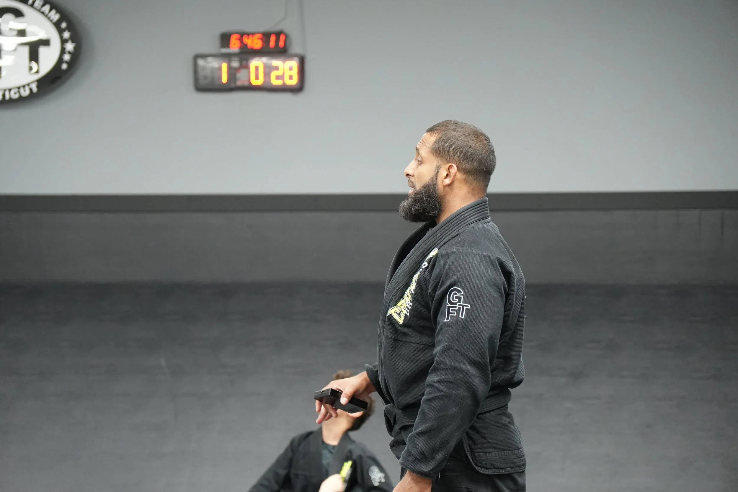 A man in a black martial arts uniform stands indoors against a gray wall with a digital clock showing 6:45:11 and a scoreboard displaying 1 and 28. He has a beard and is looking to his right, holding a black object in his right hand.