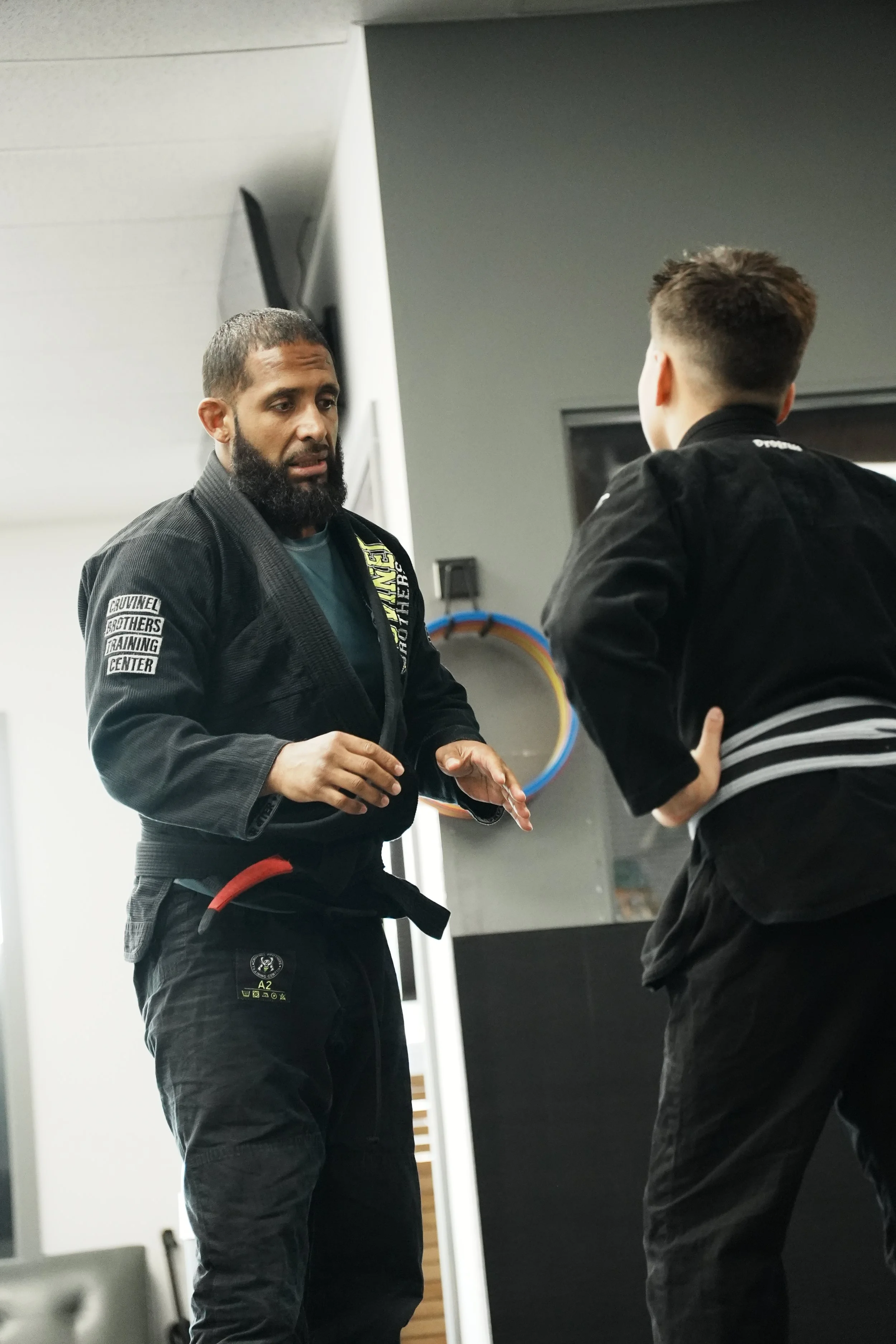 Two men in Brazilian Jiu-Jitsu gi uniforms talking inside a training facility. One man has a beard and short hair, and the other has a short haircut and is facing away from the camera.
