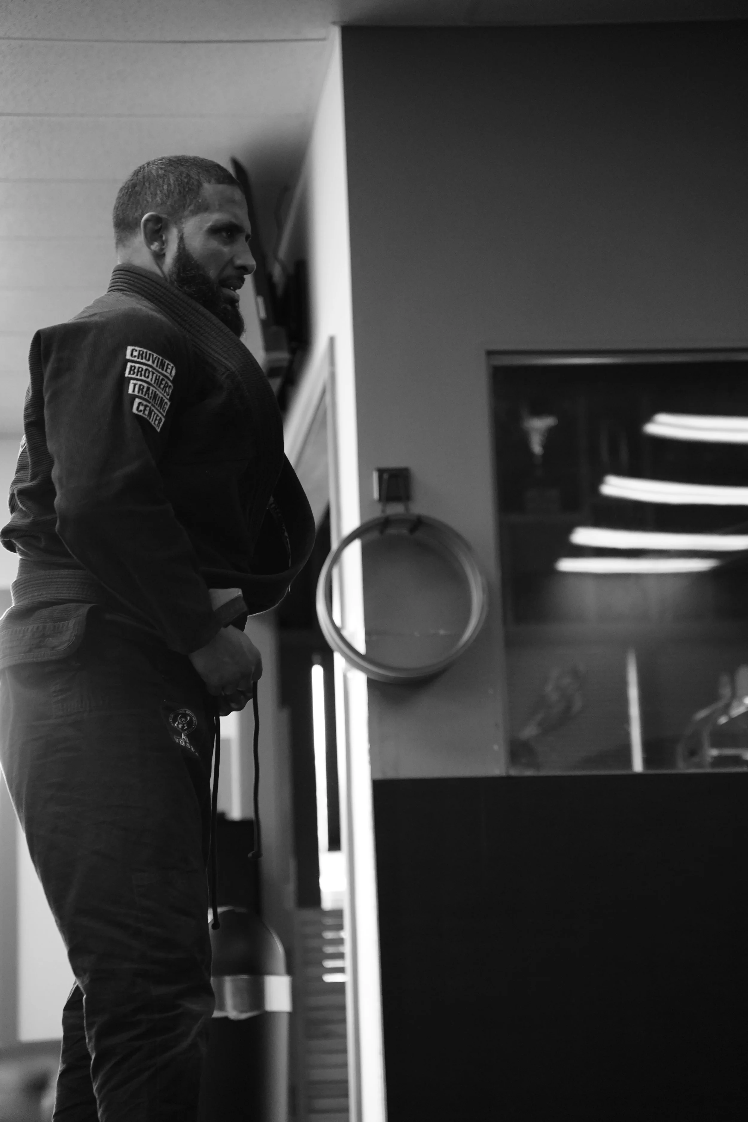 A man in a martial arts gi standing indoors, holding a belt in his hand, with a focused expression. The gi has patches on the sleeve, and the background includes a window and some shelves.