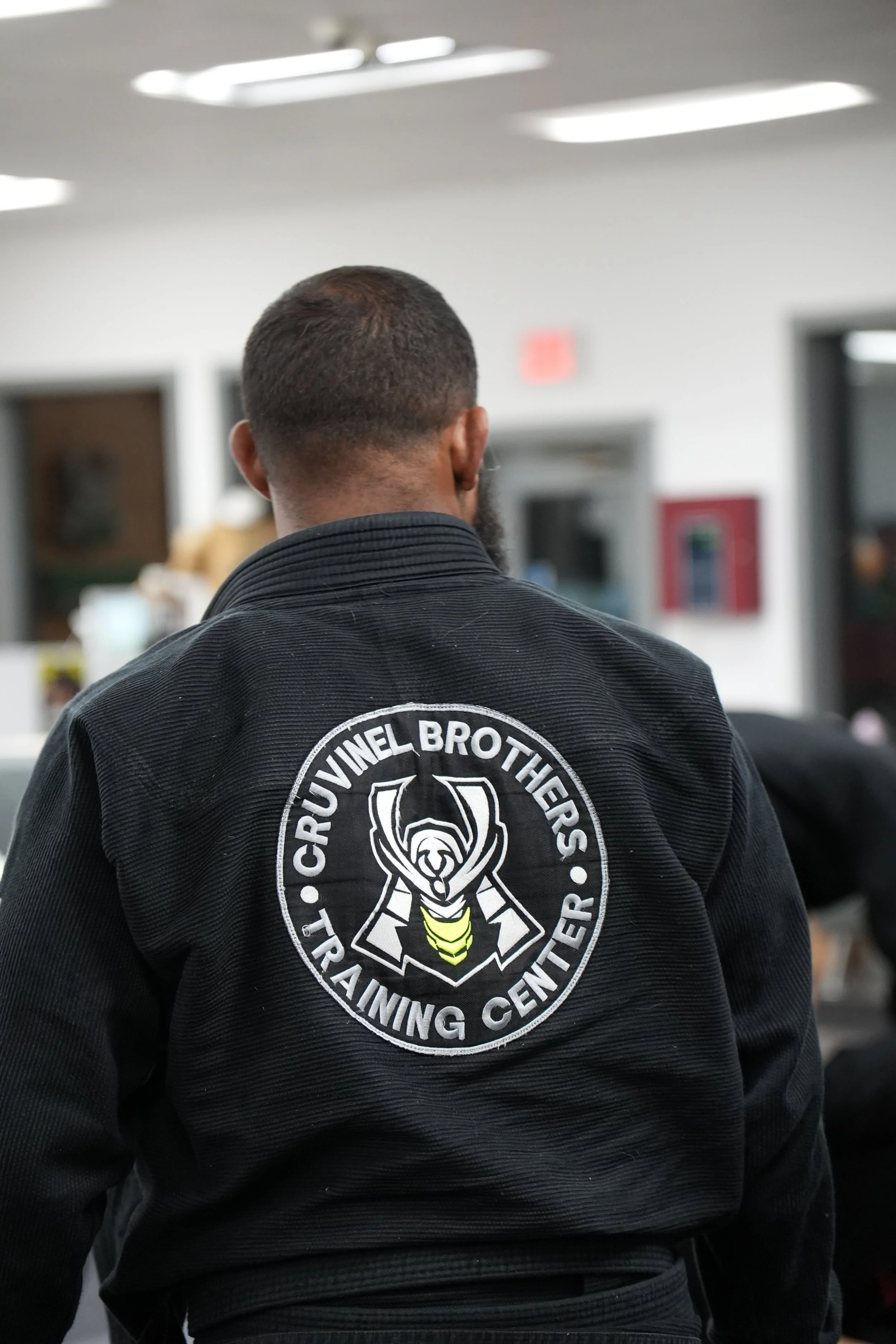 Back of a person wearing a black martial arts gi with a Patch of 'Crucivinel Brothers Training Center' on the back, standing indoors in a training facility.