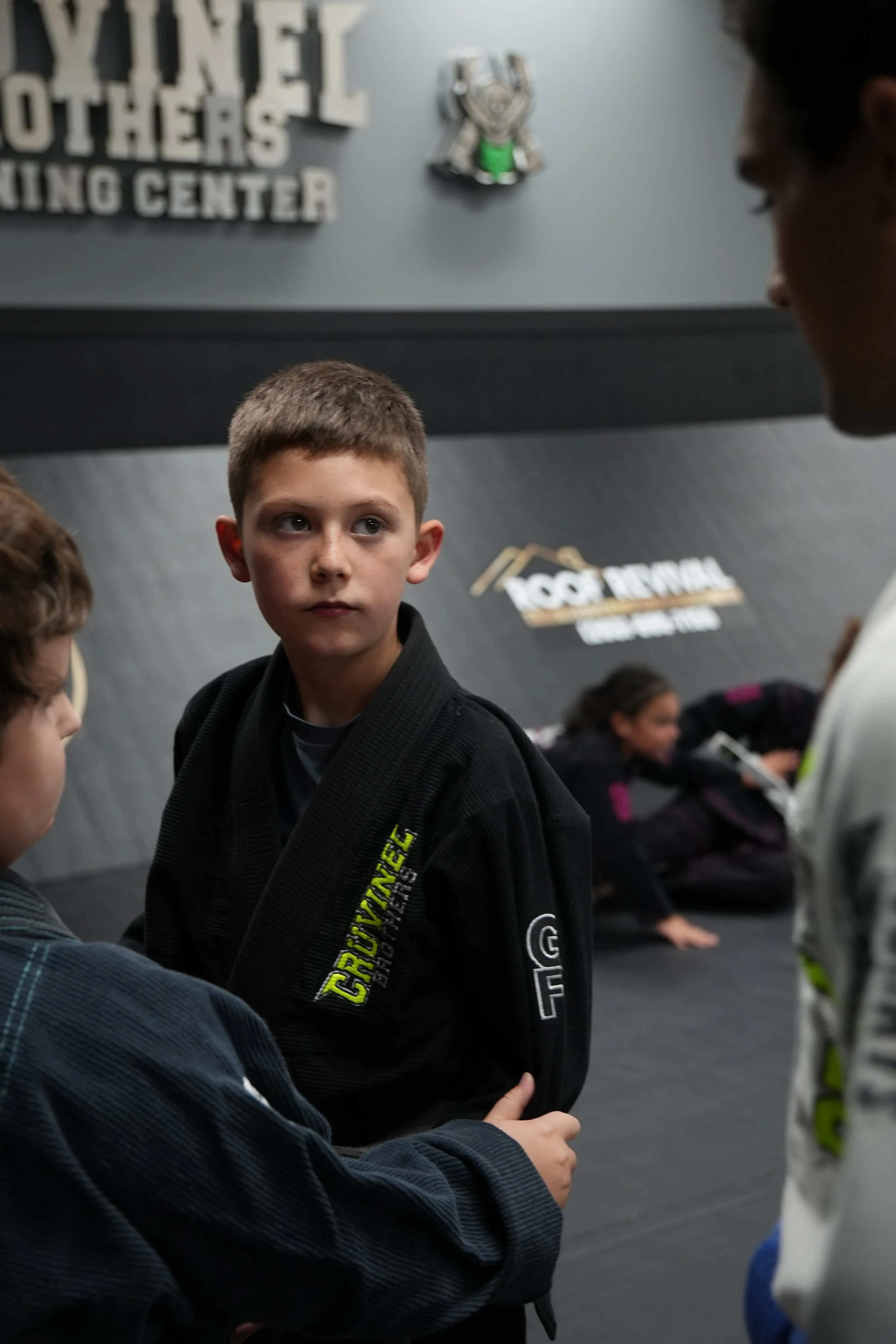 A young boy in a black martial arts gi looking towards a person in front of him, with other children in the background at a martial arts training center.