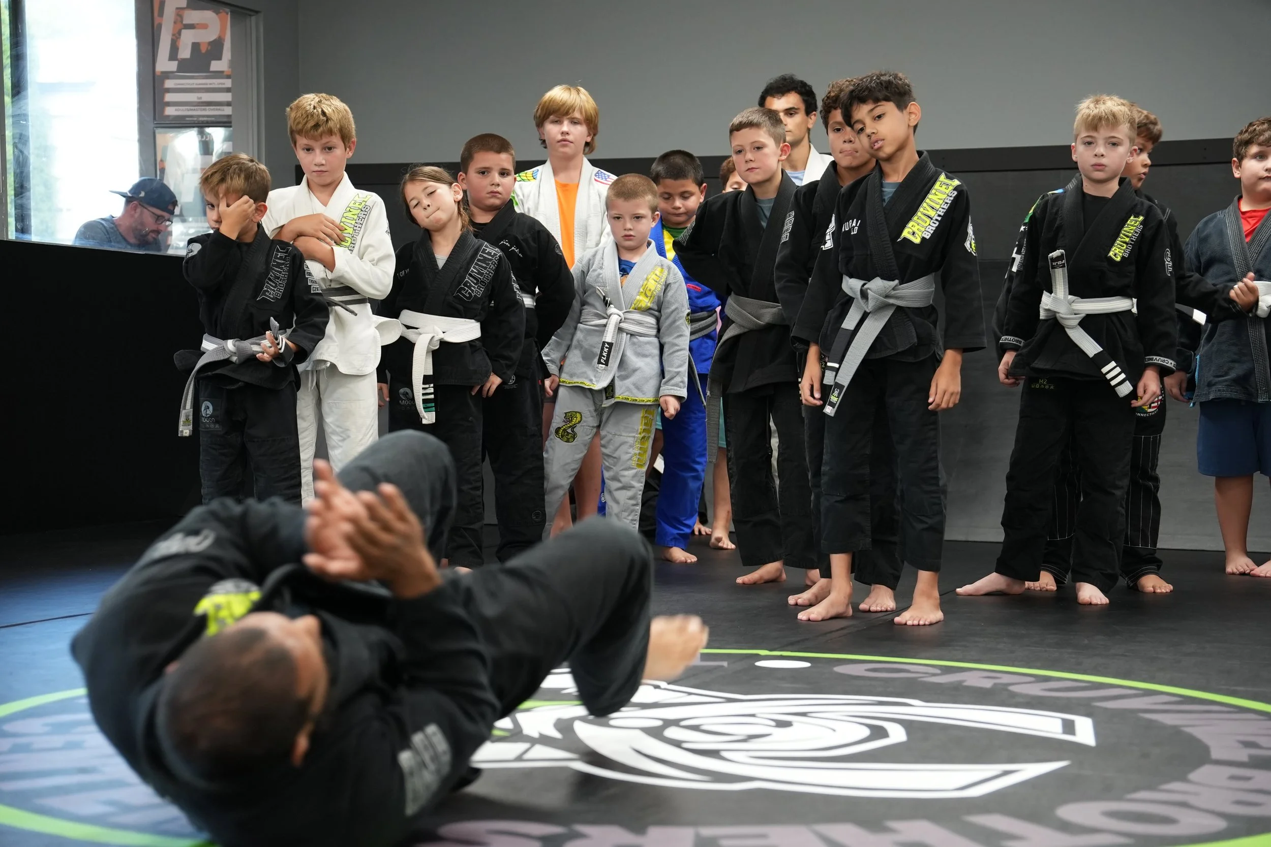 A group of children in martial arts uniforms standing in a line inside a martial arts studio, some with serious expressions, while a person in the foreground is lying on the mat. The children are wearing black, white, and gray uniforms with various p