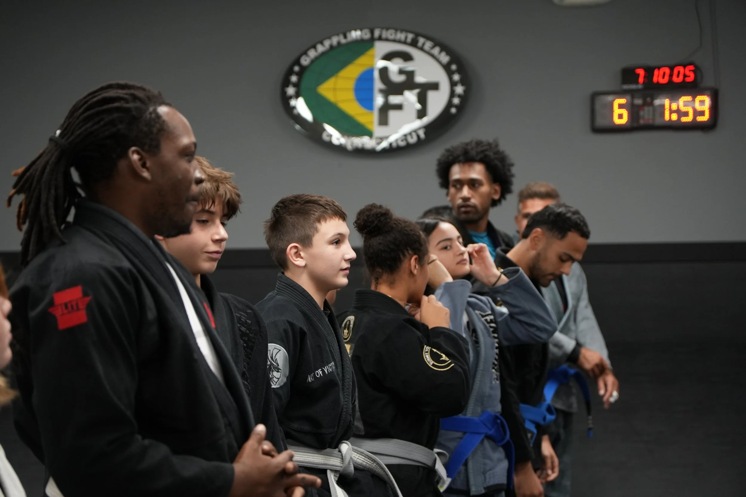 A group of diverse martial artists, including children and adults, standing in a row with their eyes closed and hands in a prayer or meditation position in a dojo or martial arts training facility. The background features a wall with a logo reading '