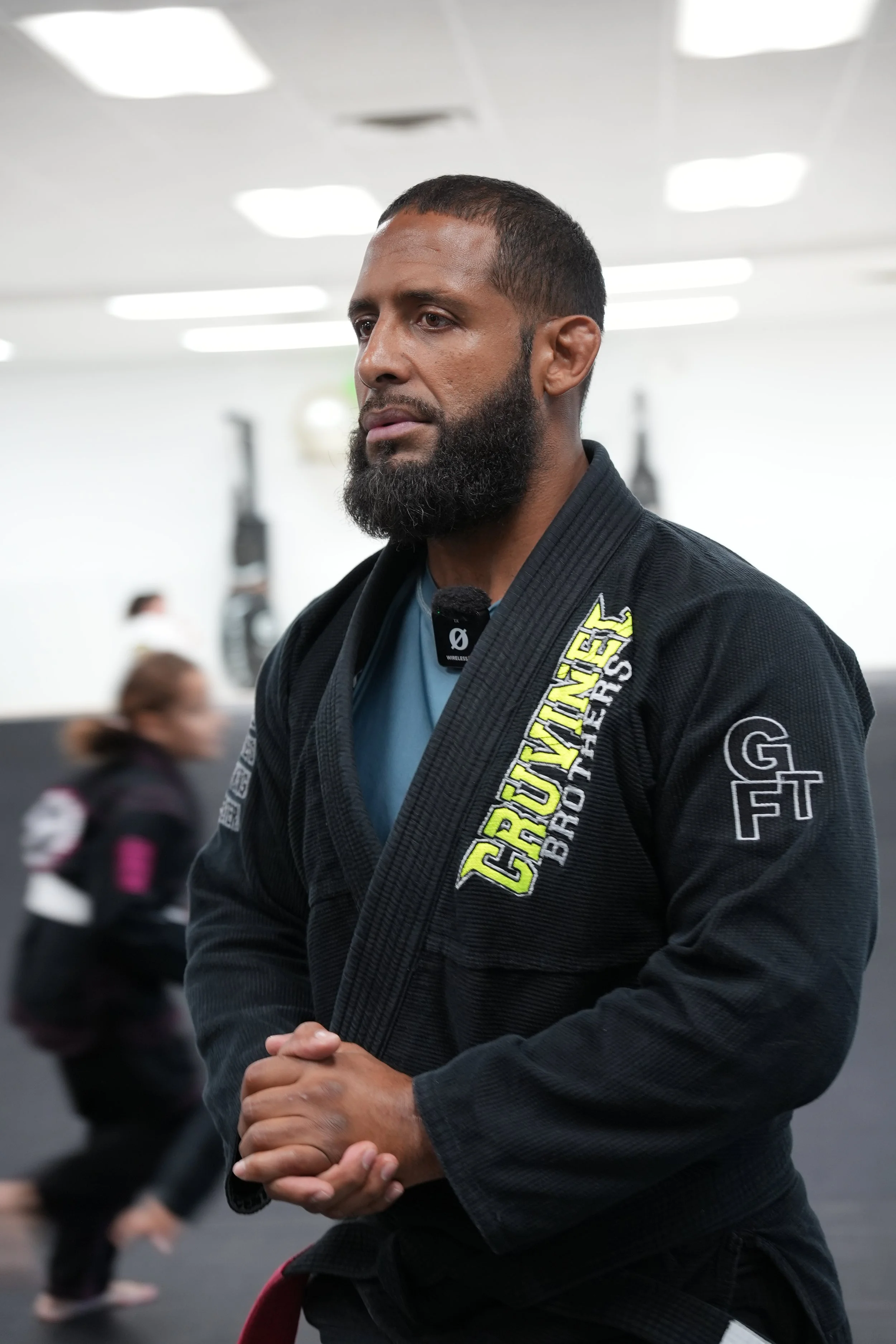 A man wearing a black Brazilian Jiu-Jitsu gi with yellow and white patches, standing with hands clasped in a martial arts training gym.