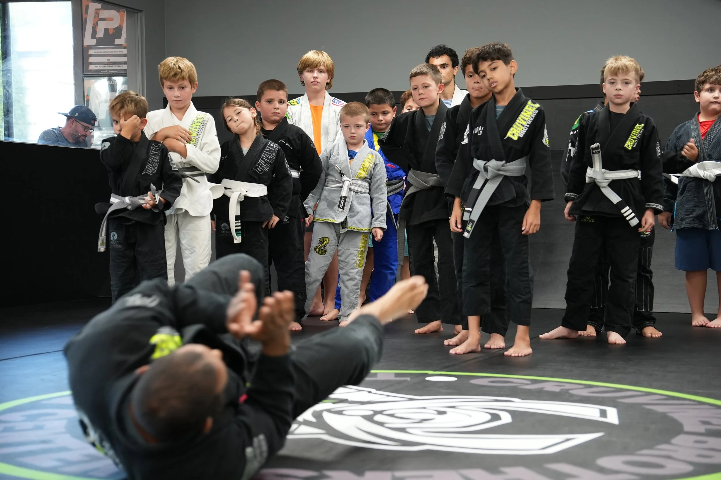 Children in martial arts uniforms standing in a line on a training mat, with some sitting or lying down in the foreground, in a martial arts gym.