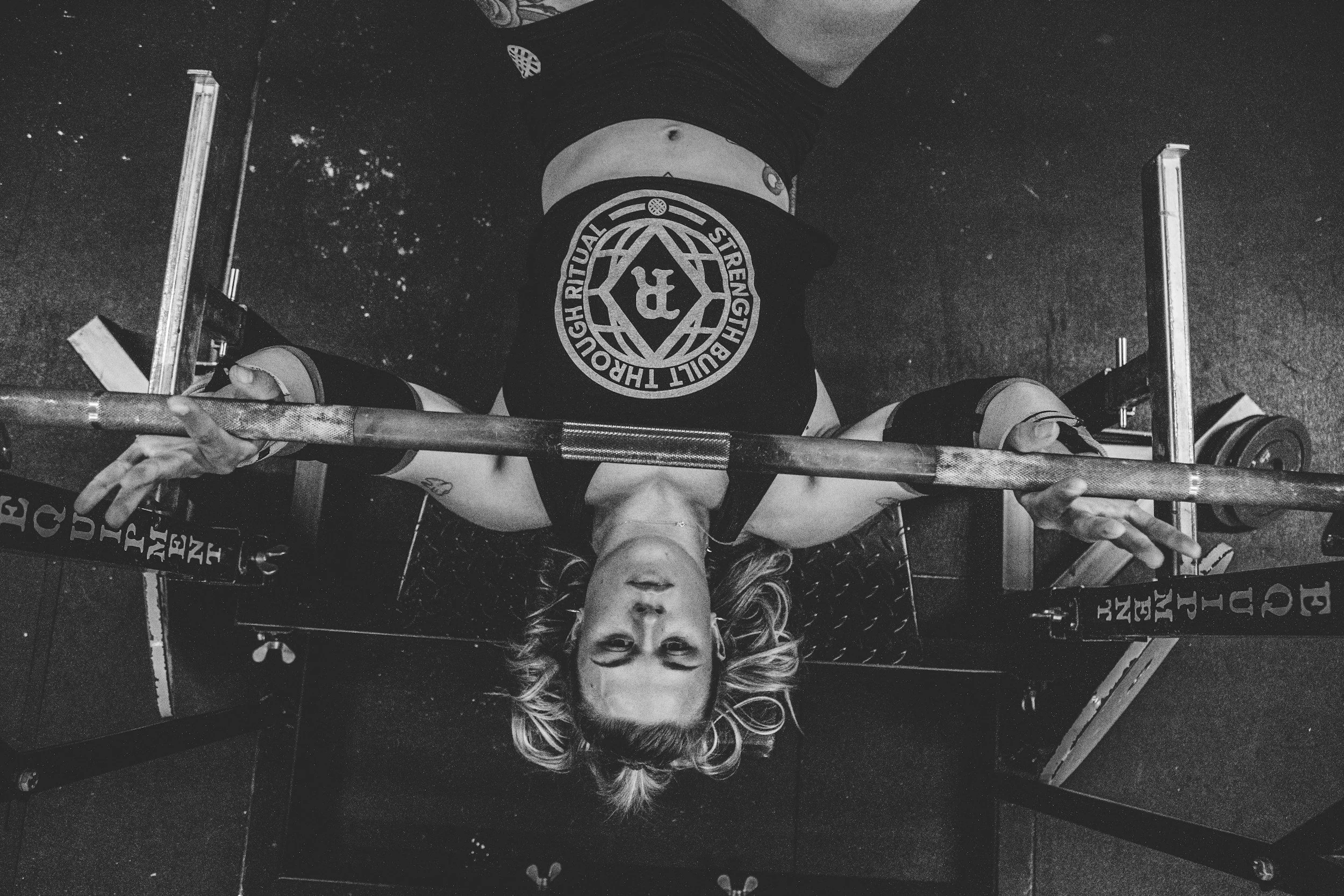 A woman with long hair doing a bench press at a gym, lifting a barbell with weights, with a determined expression on her face.
