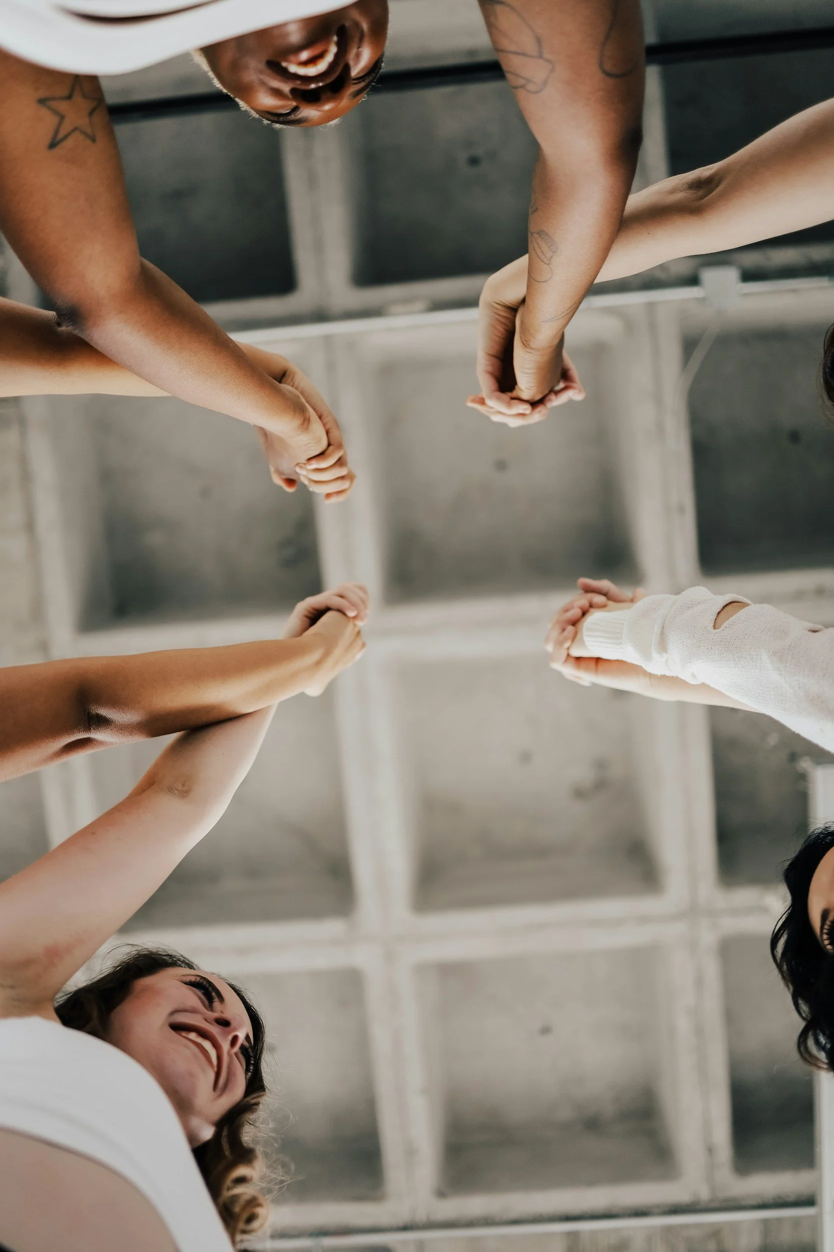 Smiling group of diverse people holding hands in a circle from below, with a ceiling grid in the background.