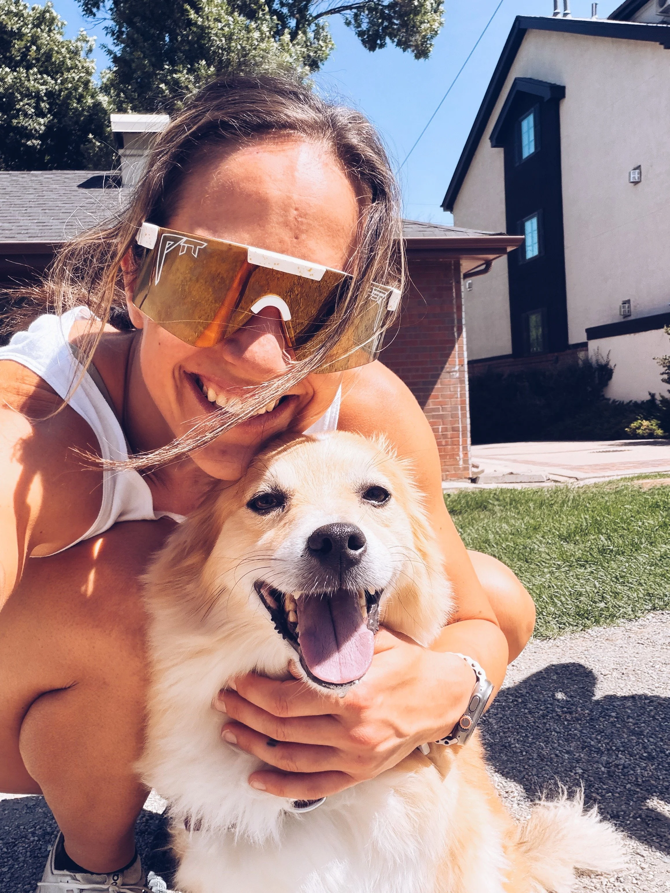 A woman with sunglasses smiling as she holds a happy, yawning, fluffy golden retriever puppy outdoors on a sunny day with houses and trees in the background.