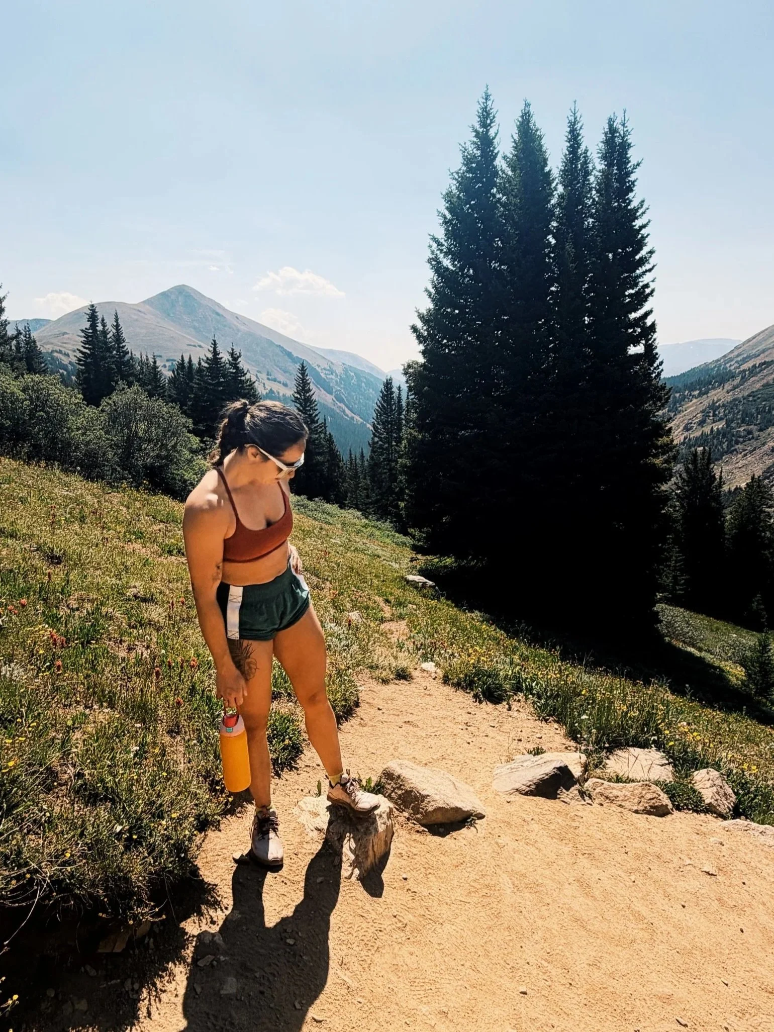 A woman wearing a sports bra, shorts, and sunglasses walking along a mountain trail surrounded by green grass and trees with mountains in the background.