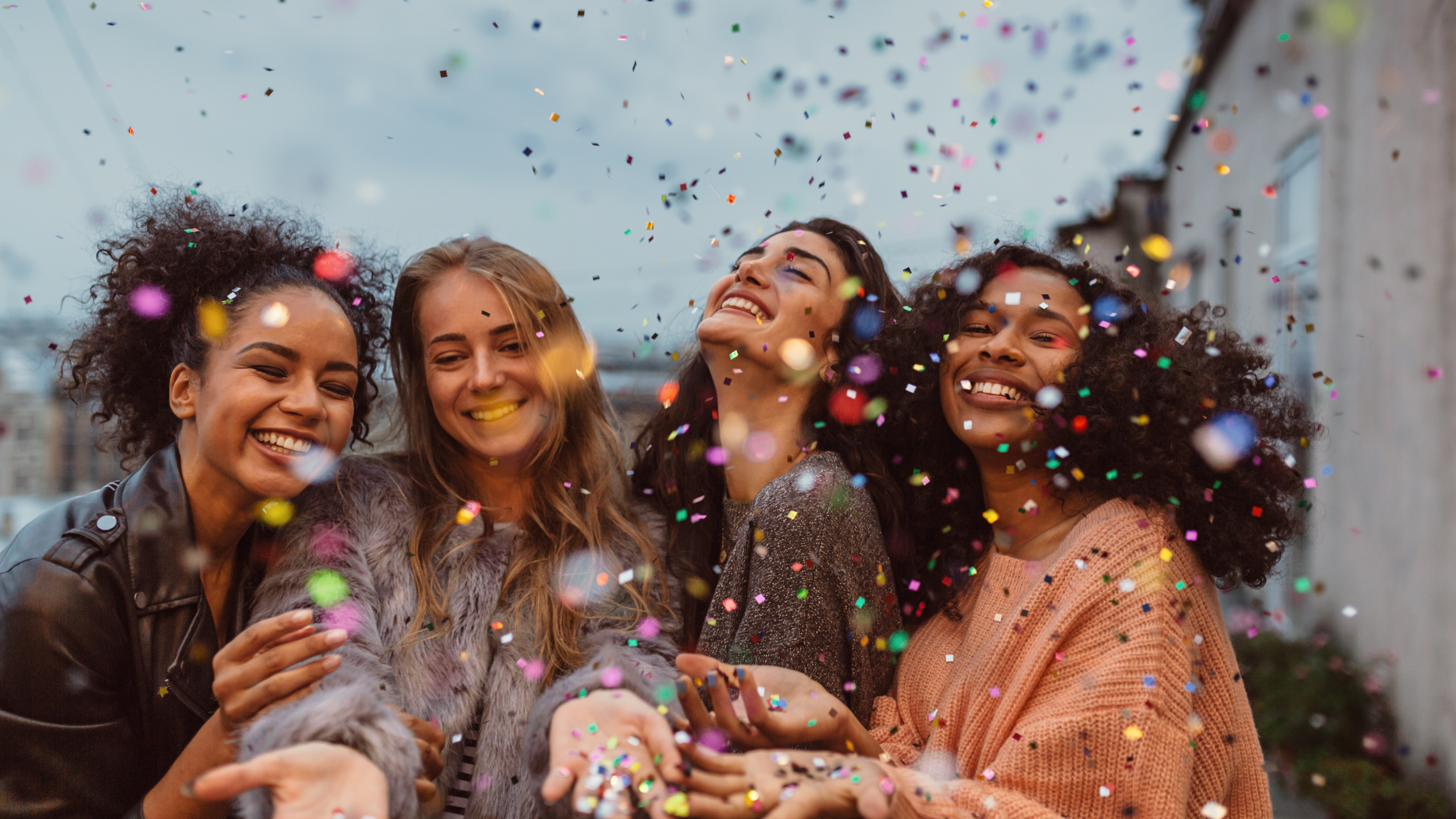Group of four women celebrating with confetti outdoors, smiling, and enjoying the moment.