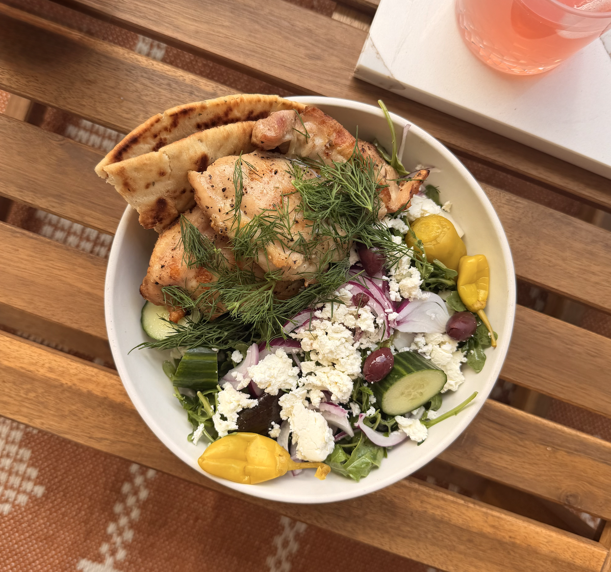 A bowl of salad with grilled chicken, pita bread, fresh vegetables, herbs, feta cheese, black olives, and yellow peppers on a wooden table.