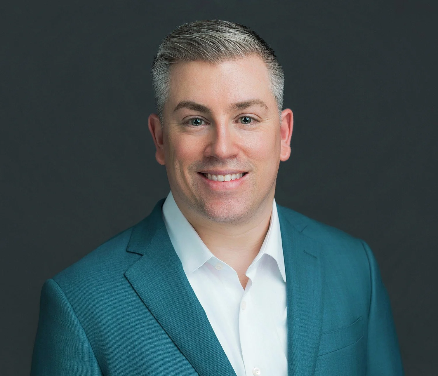 Headshot of a smiling man with gray hair, wearing a teal suit jacket and white shirt, against a dark gray background.