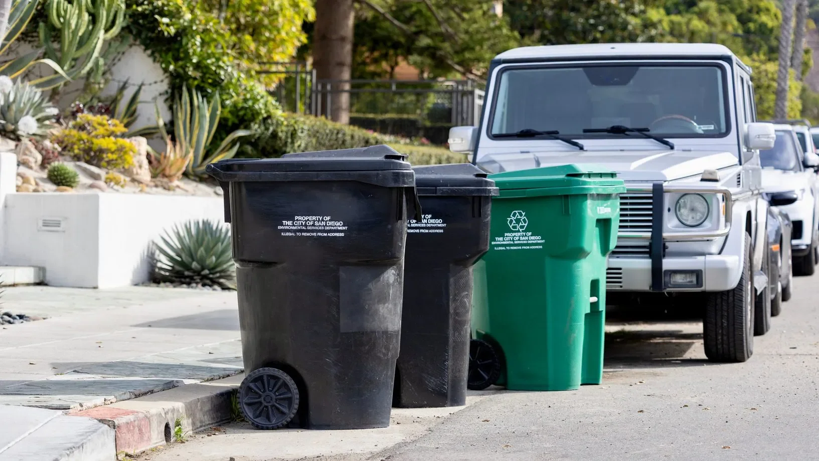 Three trash bins, two black and one green, lined up on a sidewalk next to a white SUV with trees and plants in the background.