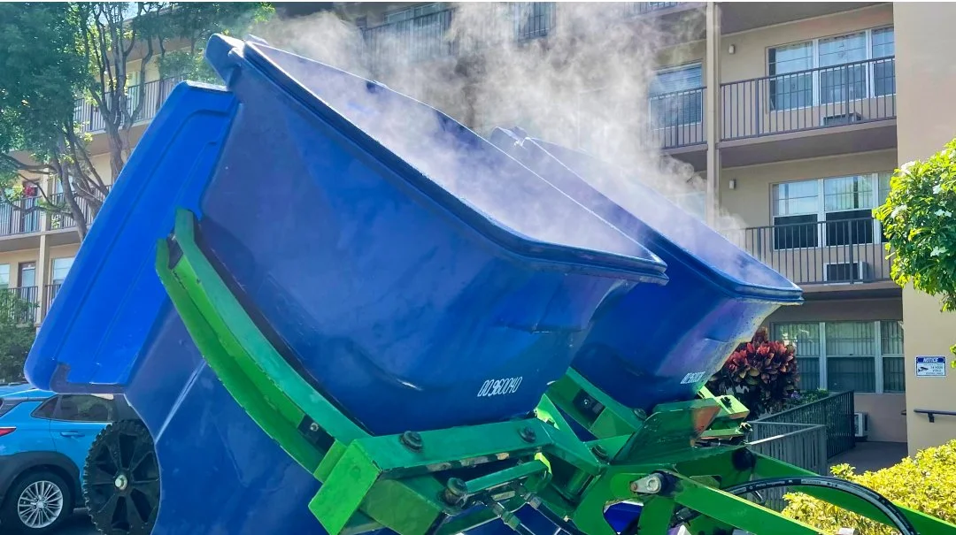 A dumpster with its lid open and steam coming out, situated outdoors in front of an apartment building, with parked cars and trees in the background.