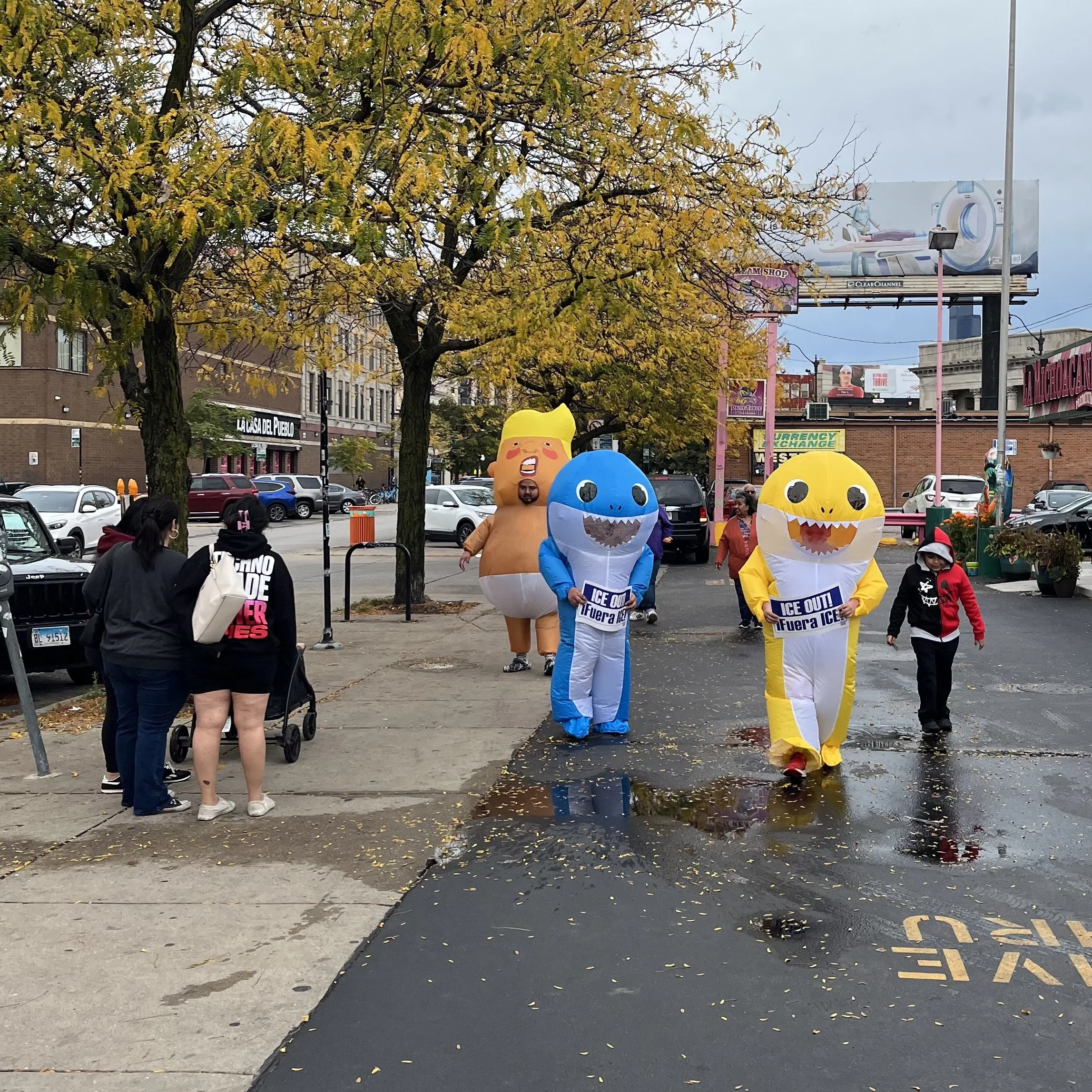 People in colorful dinosaur costumes holding signs that say "Ice Out! Fuera ICE!" walking on a city street with trees, parked cars, and billboards in the background.