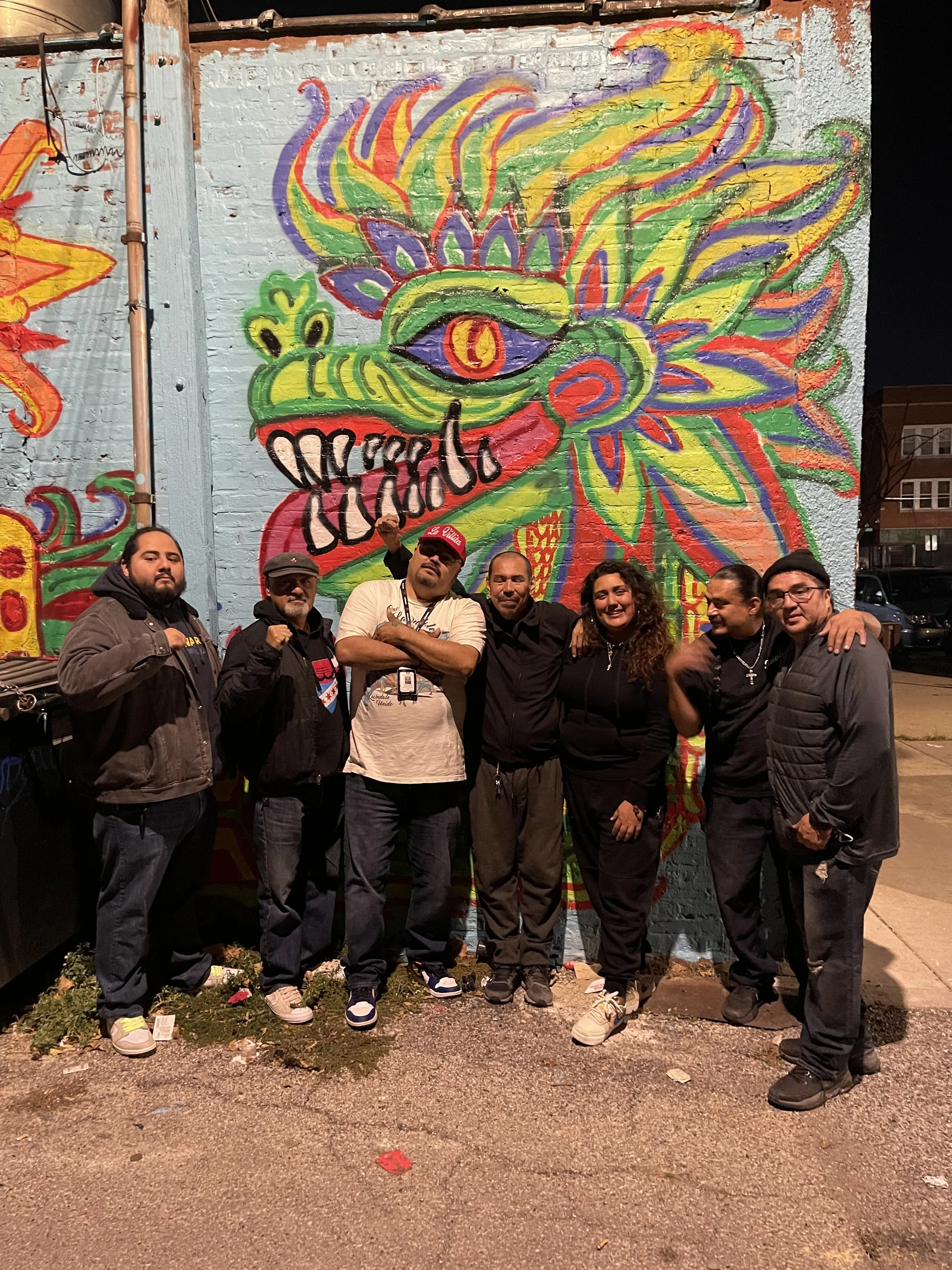 A group of six people standing in front of a colorful mural of a stylized dragon with an eye, on a brick wall at night. The mural features bright colors like yellow, green, purple, red, and orange. The group is posing and smiling, some with fists rai