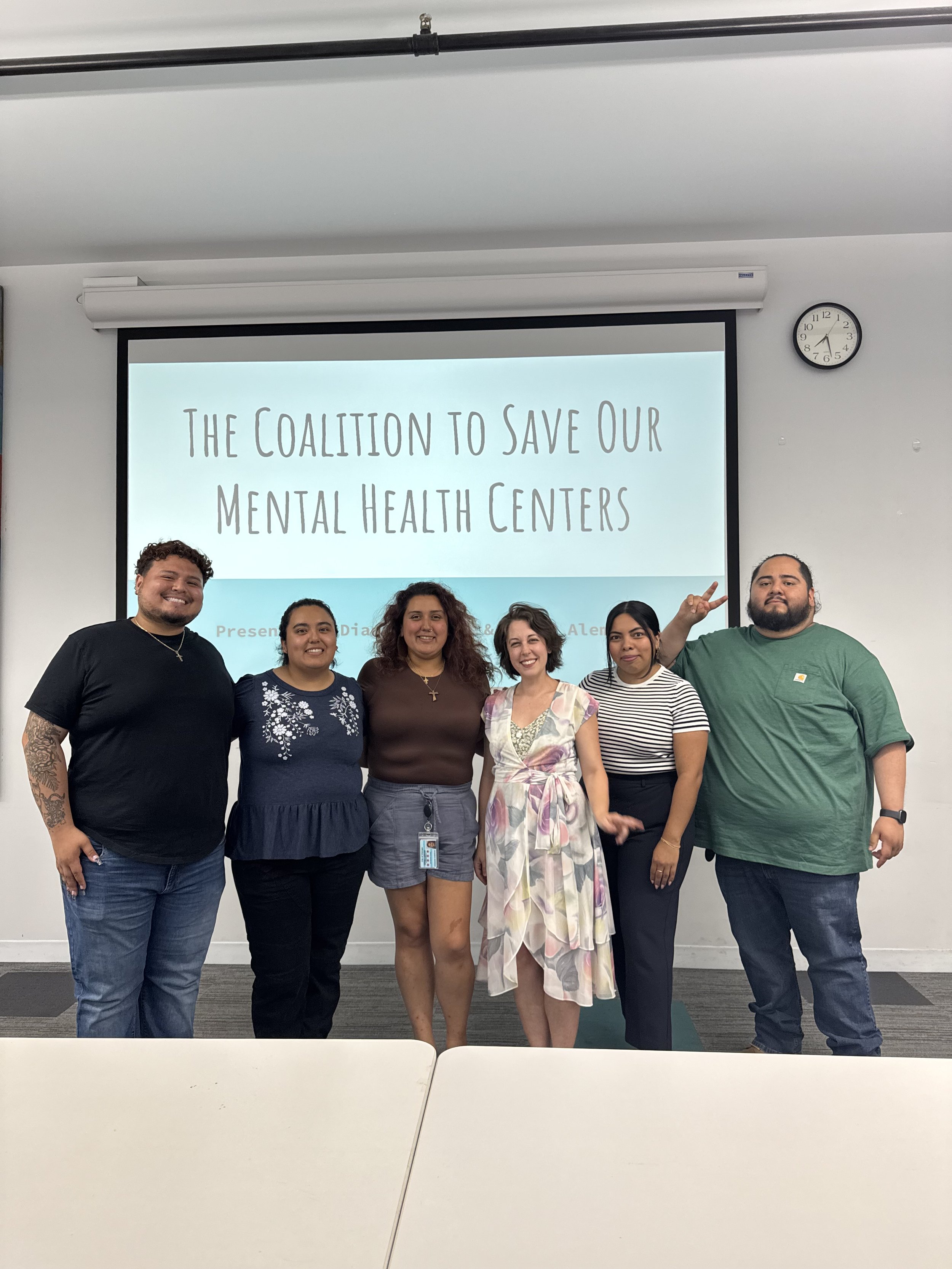 A group of seven diverse people standing in front of a projected presentation that reads 'The Coalition to Save Our Mental Health Centers,' smiling and posing for the camera.