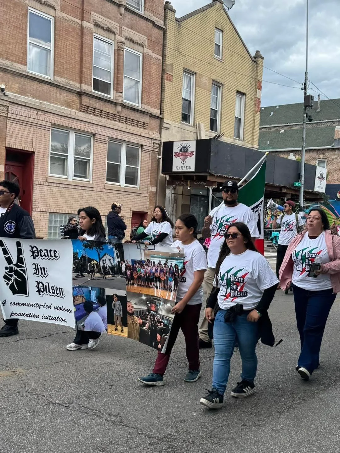 People participating in a parade march, holding a banner that reads 'Peace In Pilsen' and features photos of community activities, with buildings and cloudy sky in the background.