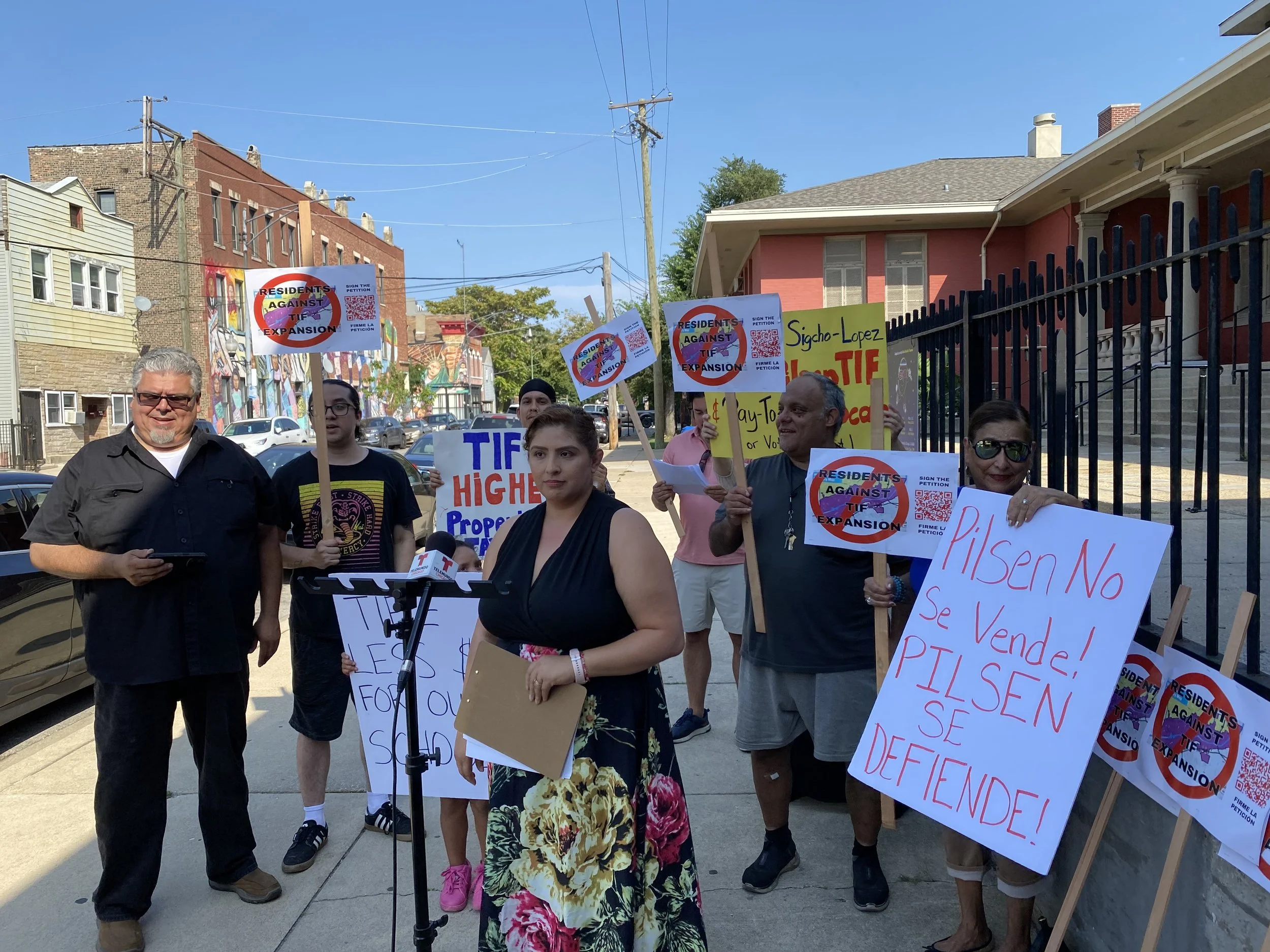 A group of people protesting on a city sidewalk holding signs against the expansion of TIF. One sign reads 'RESIDENTS AGAINST TIF EXPANSION' with a QR code. Other signs include 'TIF HIGHER Property' and messages in Spanish. Some individuals are speak