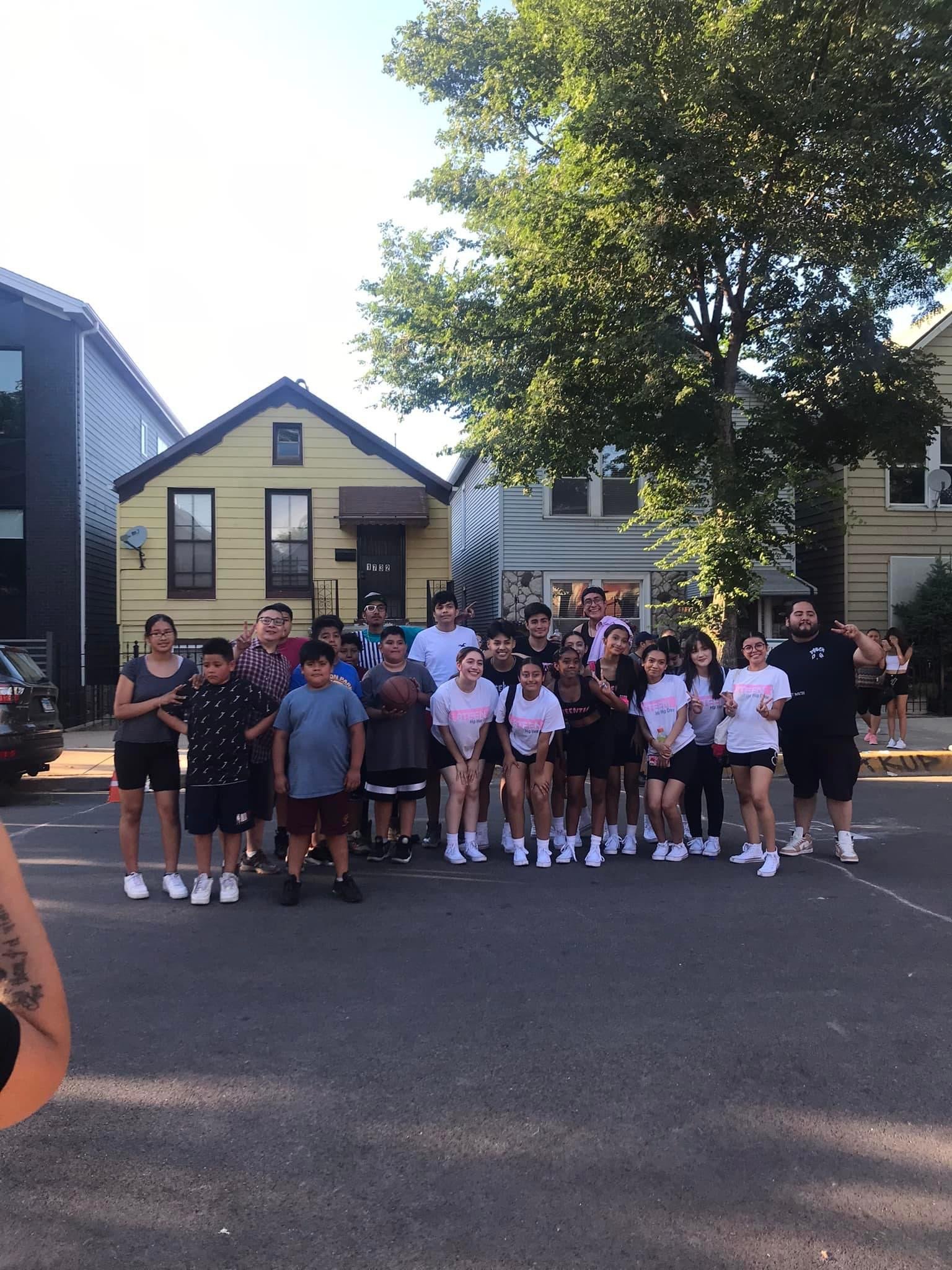 A group of young people and adults standing outside in front of houses, posing for a photo on a sunny day.