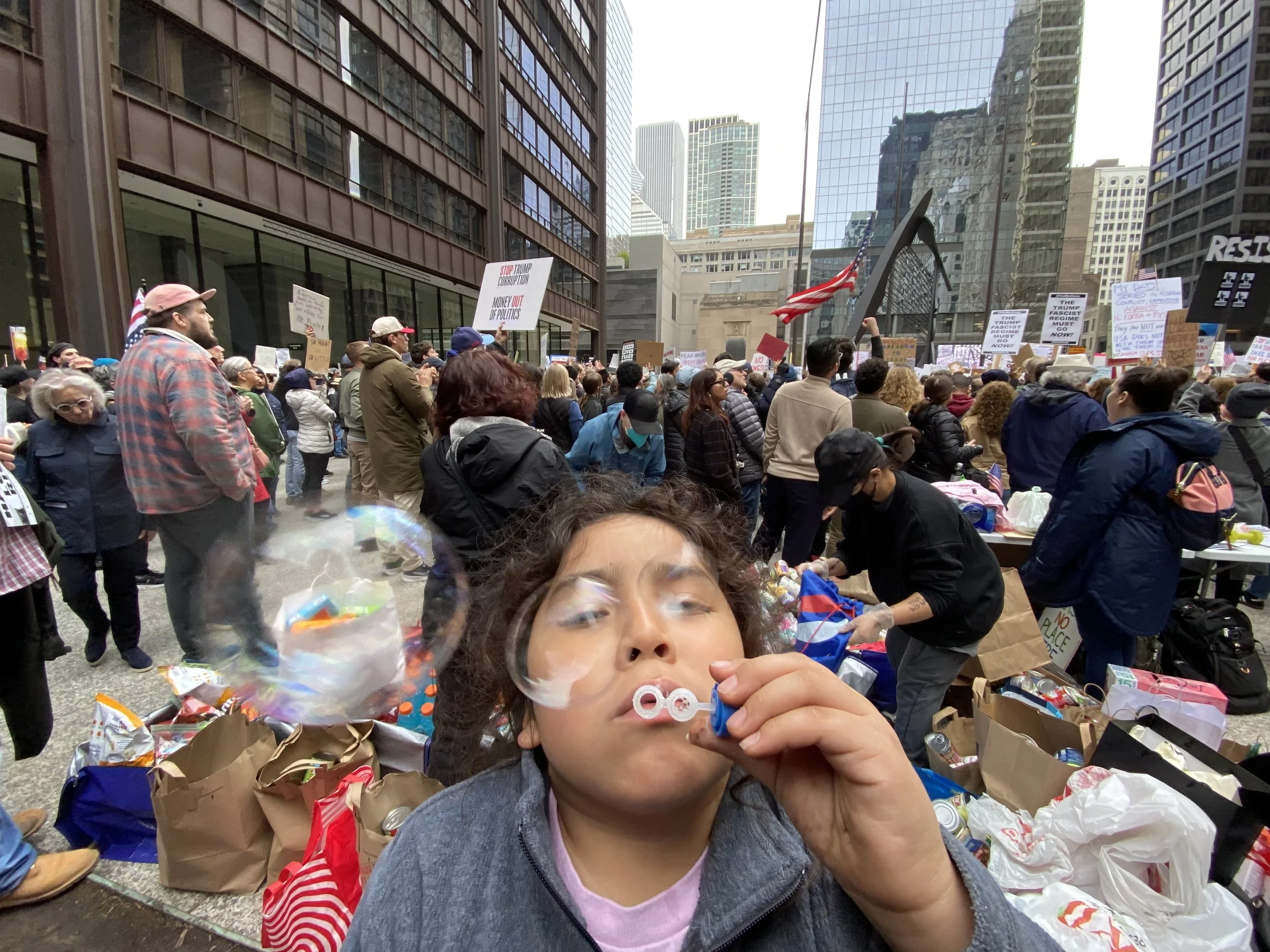 A young girl with glasses holding a bubble blower and bubbles in an urban protest or rally setting, with many people holding signs and American flags, surrounded by tall buildings.