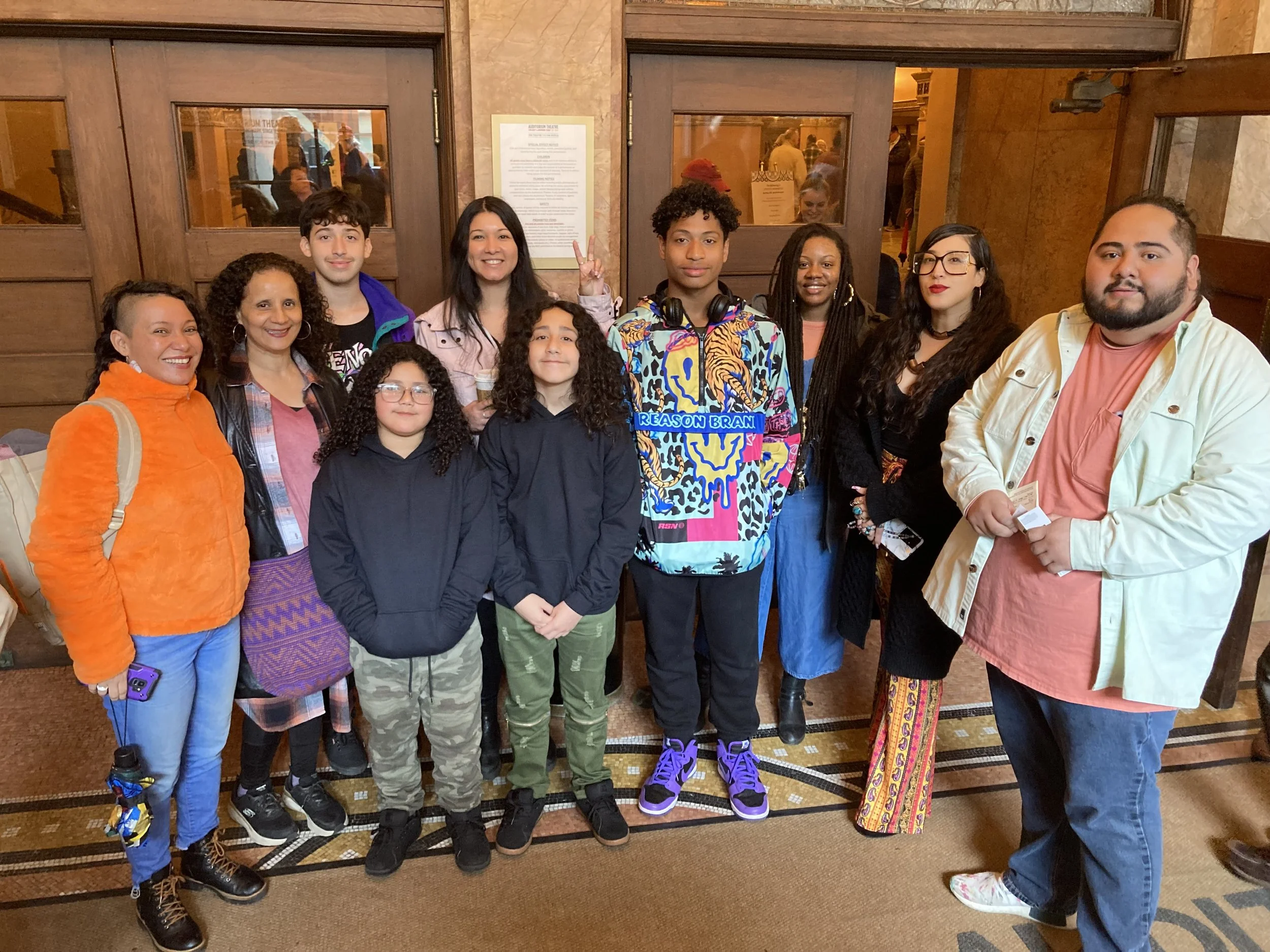 Group of nine people, including children and adults, standing in a lobby area with wooden doors and a sign on the wall behind them. They are smiling and dressed casually.
