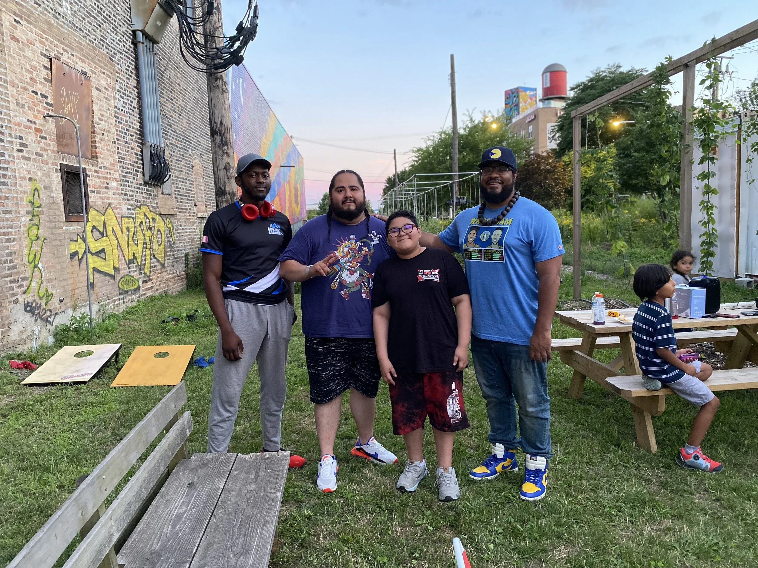 A group of four boys standing outdoors in front of a brick wall decorated with colorful graffiti art, with greenery and additional children sitting at picnic tables nearby. One boy is making a peace sign.