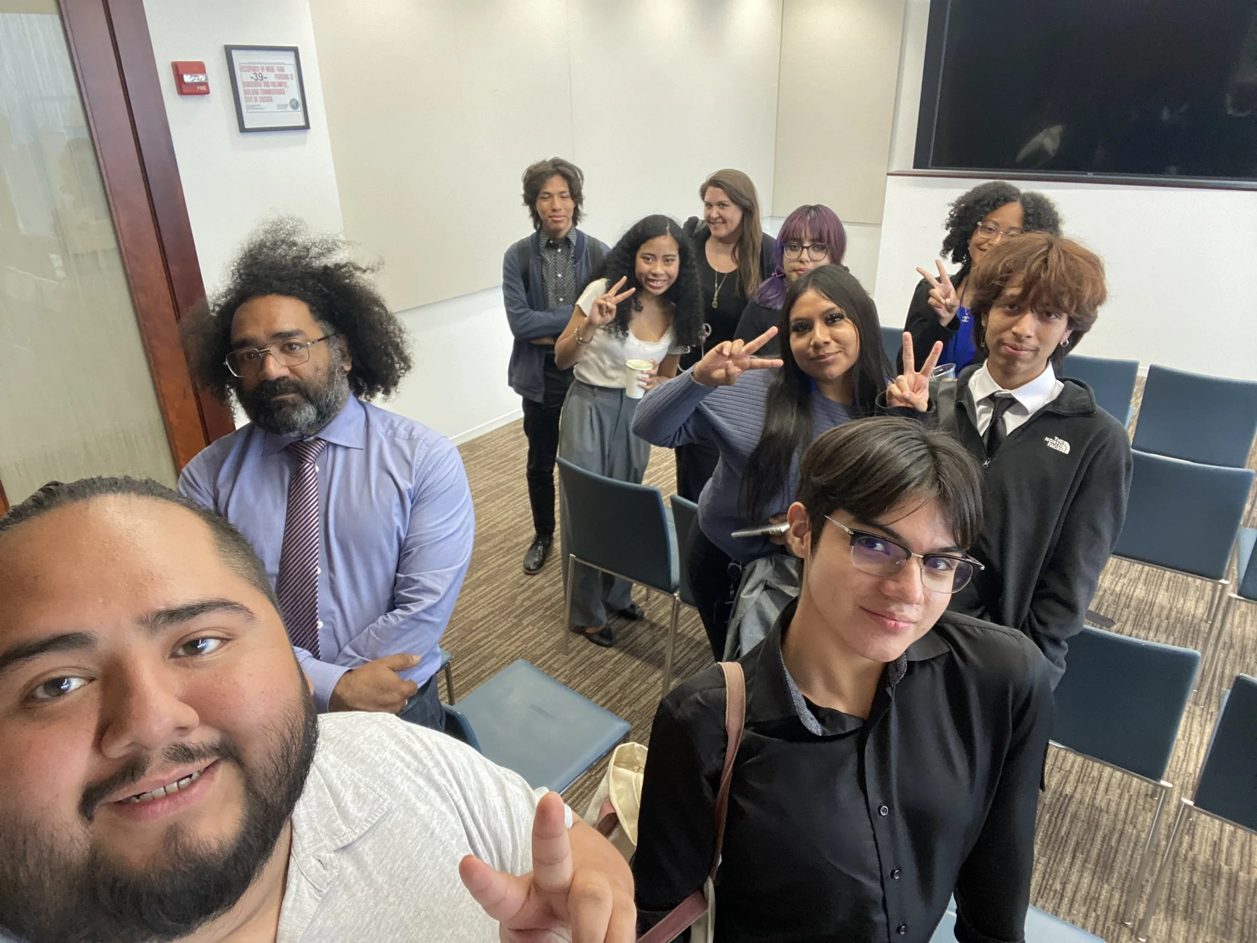 Group of diverse young adults and one older man taking a selfie together in a conference room, some making peace signs, with a large black TV screen and wall decorations visible in the background.