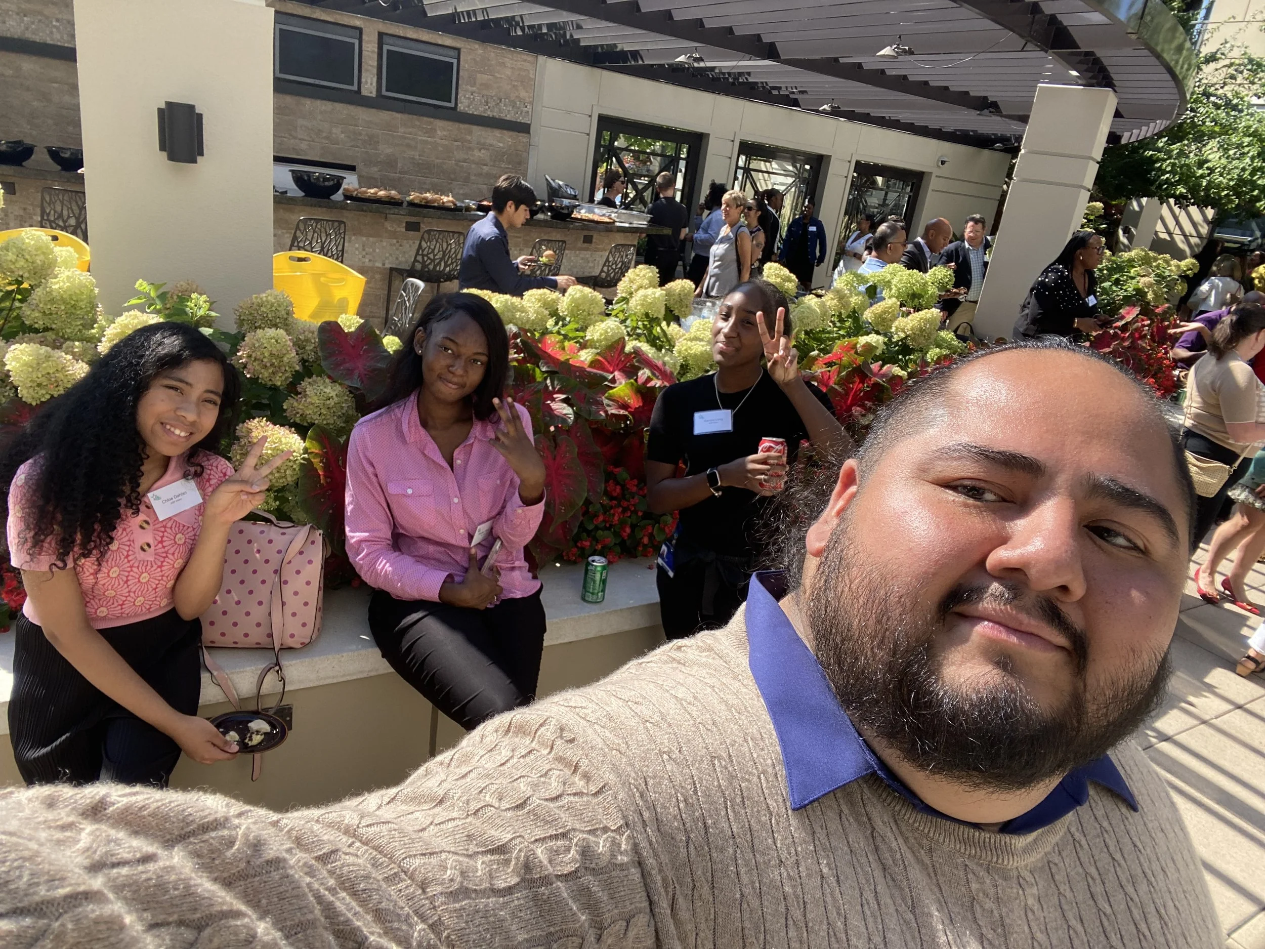 A group of people gathered outdoors at a social event. Two young women sit in the front, both making peace signs with their hands, while the woman on the left holds a plate of food and a pink purse. The crowd in the background is mingling around a bu