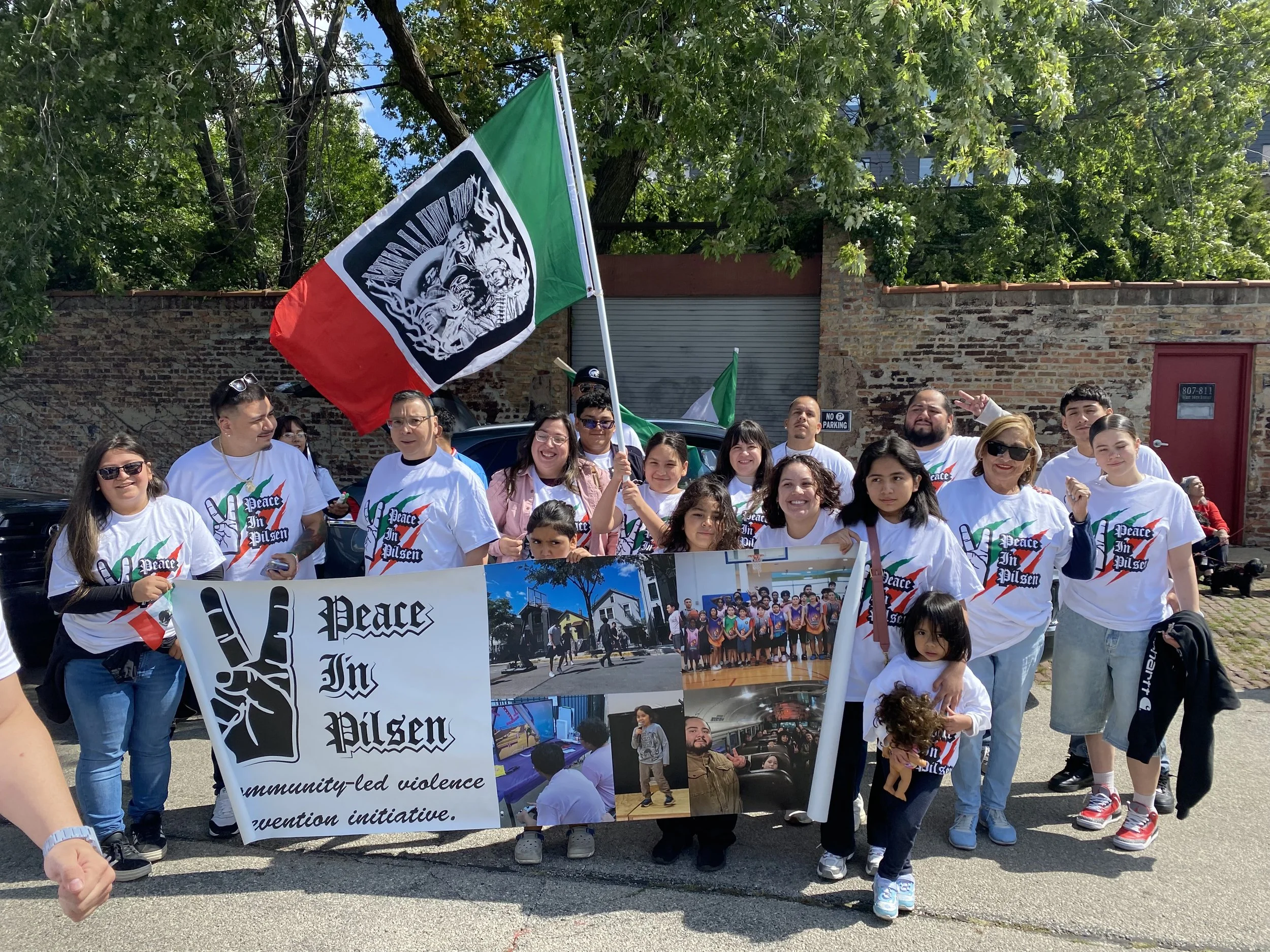 A diverse group of people participating in a community march or rally, holding banners and flags, with large trees and a brick wall in the background.