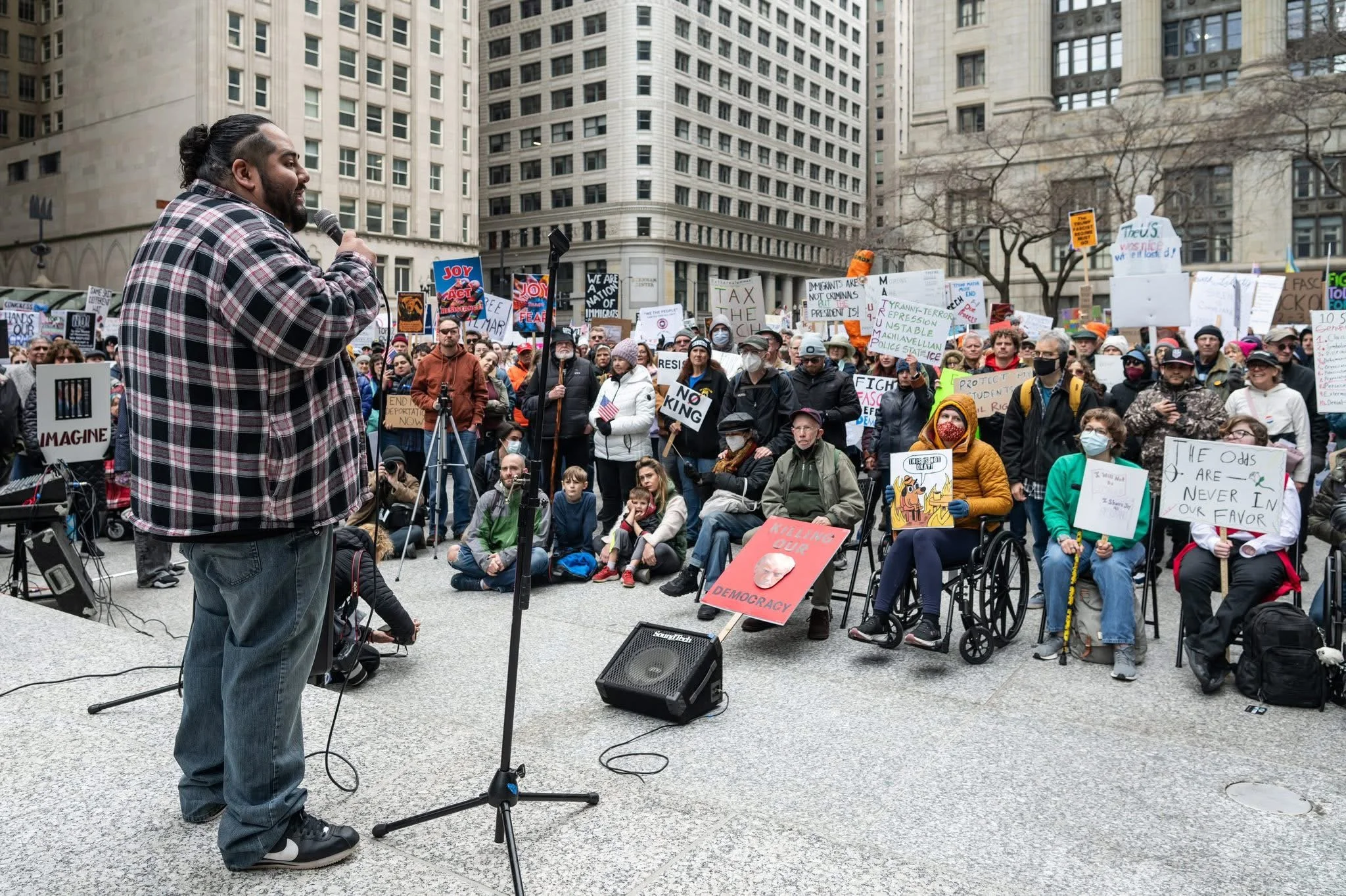 Leo, with long dark hair tied back, wearing a plaid shirt and jeans, speaking into a microphone at an outdoor protest gathering.