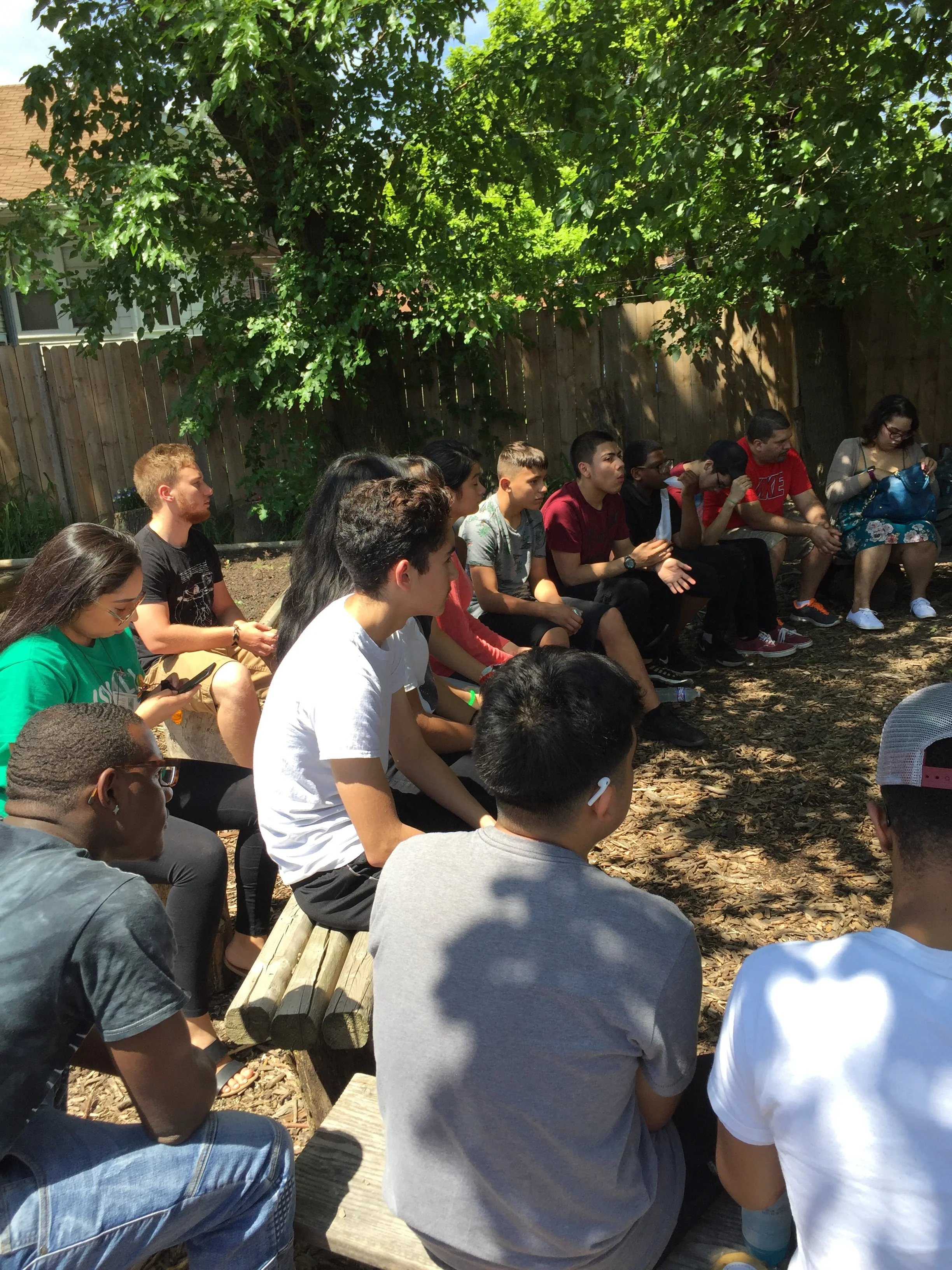 A group of diverse young people sitting outdoors on wooden benches under leafy trees in a backyard. They seem to be listening attentively to someone or something, with some looking ahead, and some using their phones.