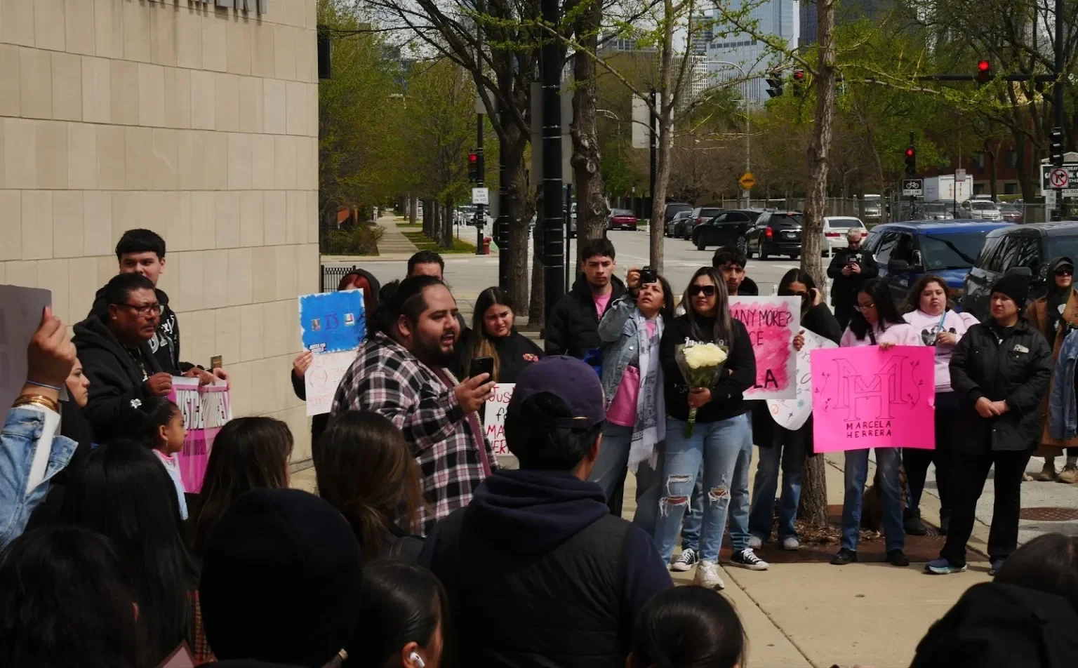People gathered outside on a sidewalk, participating in a protest or rally. Some are holding signs, including one pink sign with a large letter 'M' and the name Marcela Herrera. A man in plaid is speaking to the crowd while holding a microphone. Seve