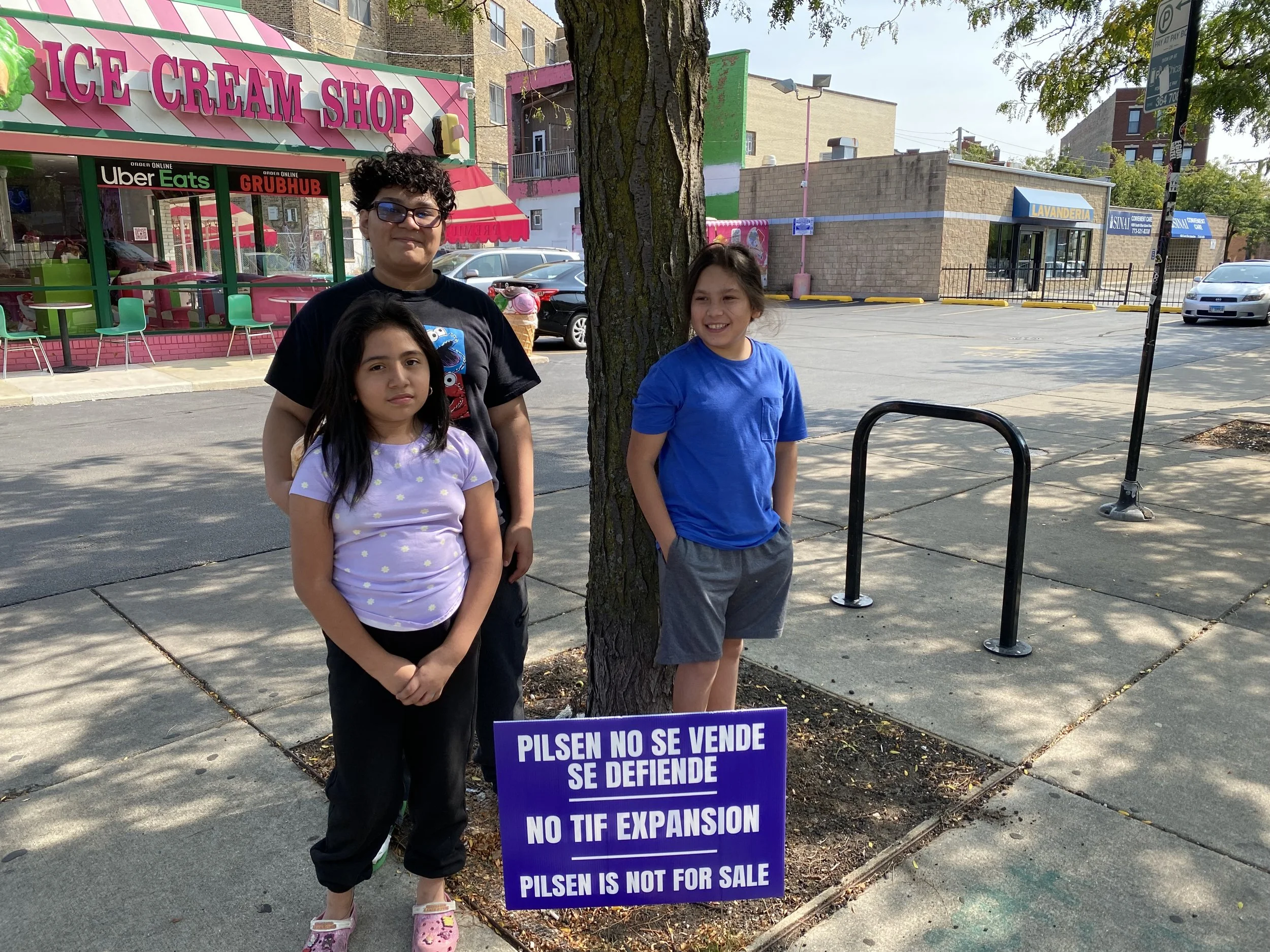 Three children standing on a sidewalk behind a sign that says, 'Pilsen no se vende, se defiende, no tiene expansión, Pilsen no for sale.' In the background, there is an ice cream shop, a corner store, parked cars, and a tree providing shade.